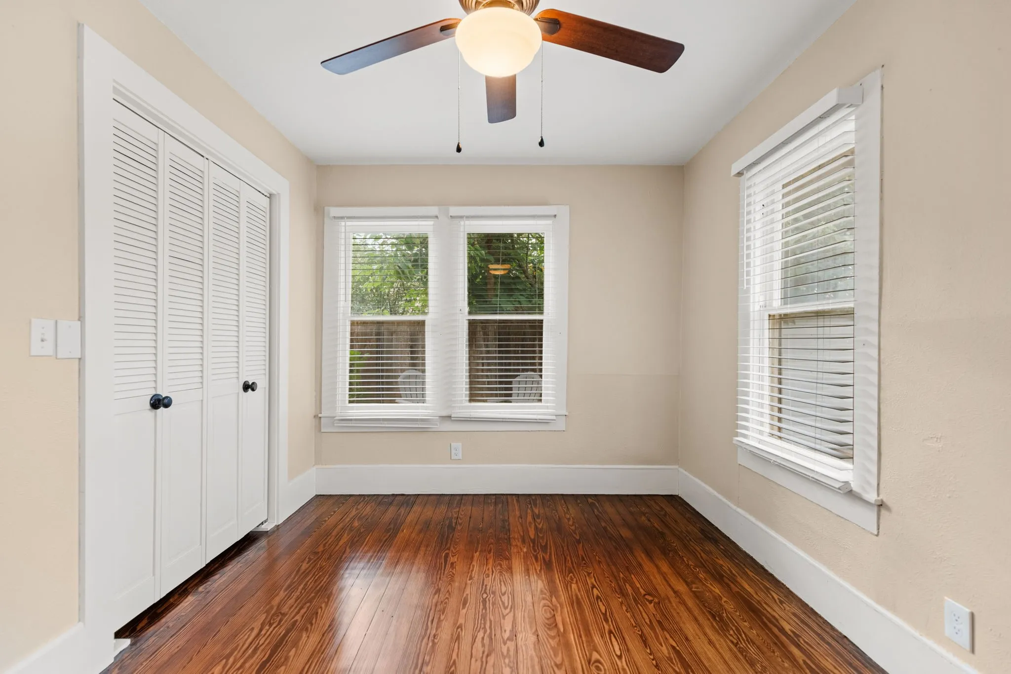 Warm-toned room featuring rich wood-finish flooring, two windows with blinds, bi-fold louvered closet doors, and a ceiling fan with light fixture