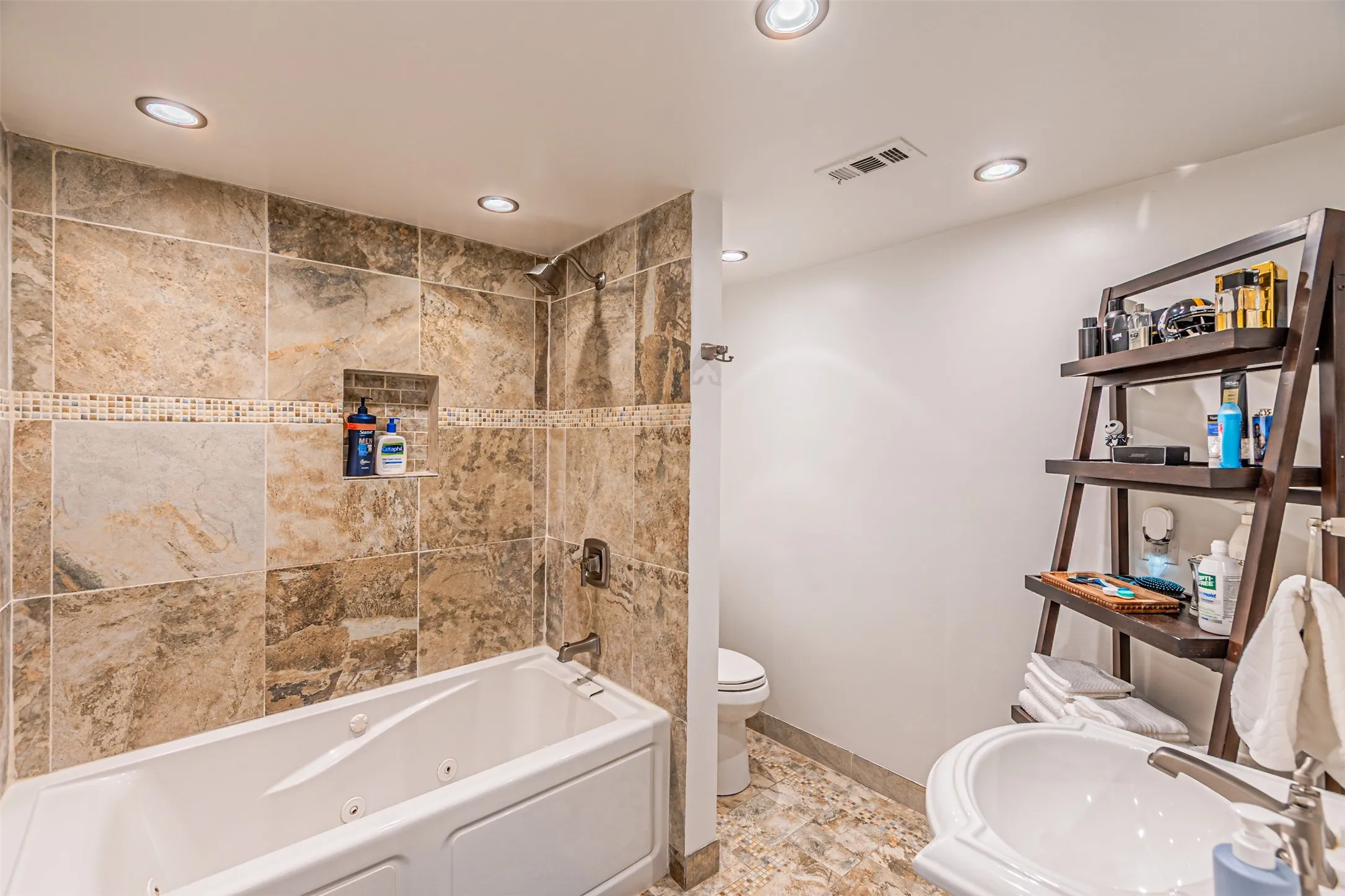 Bathroom featuring a white jetted tub, stone-finish wall and floor tiling, recessed lighting, and a pedestal sink with a contemporary faucet