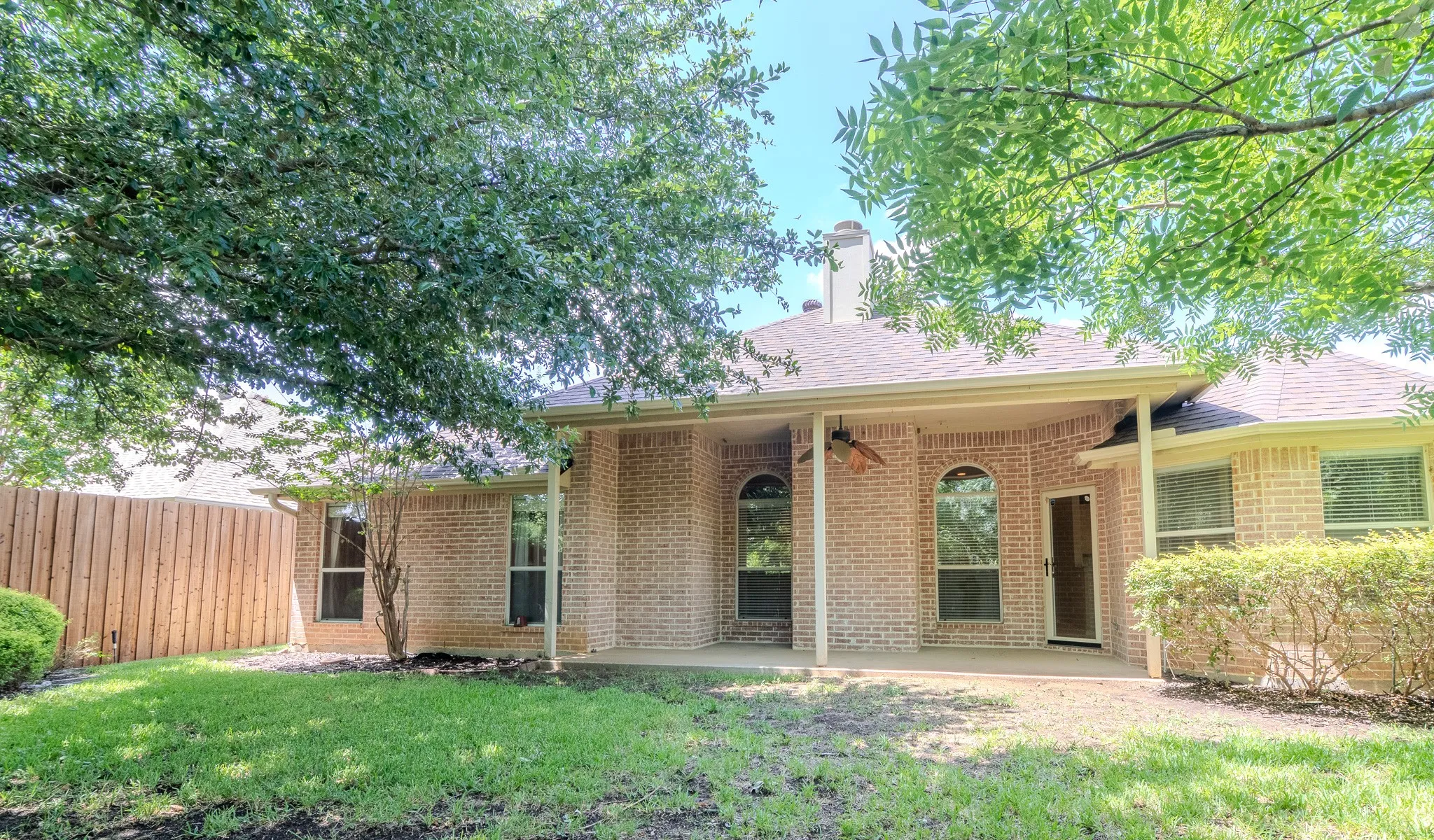 Expansive backyard featuring a covered patio, brick exterior, arched windows, privacy fencing, and mature trees