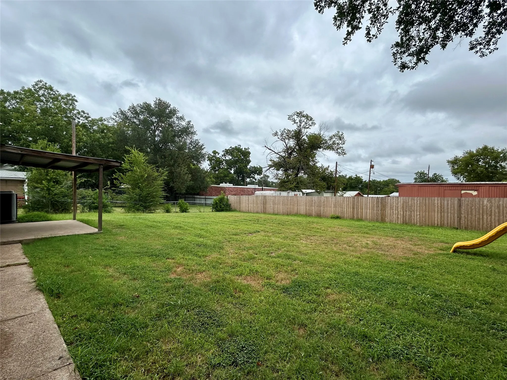 Expansive backyard featuring a covered patio, concrete walkway, full perimeter fencing, mature trees, and a spacious lawn area