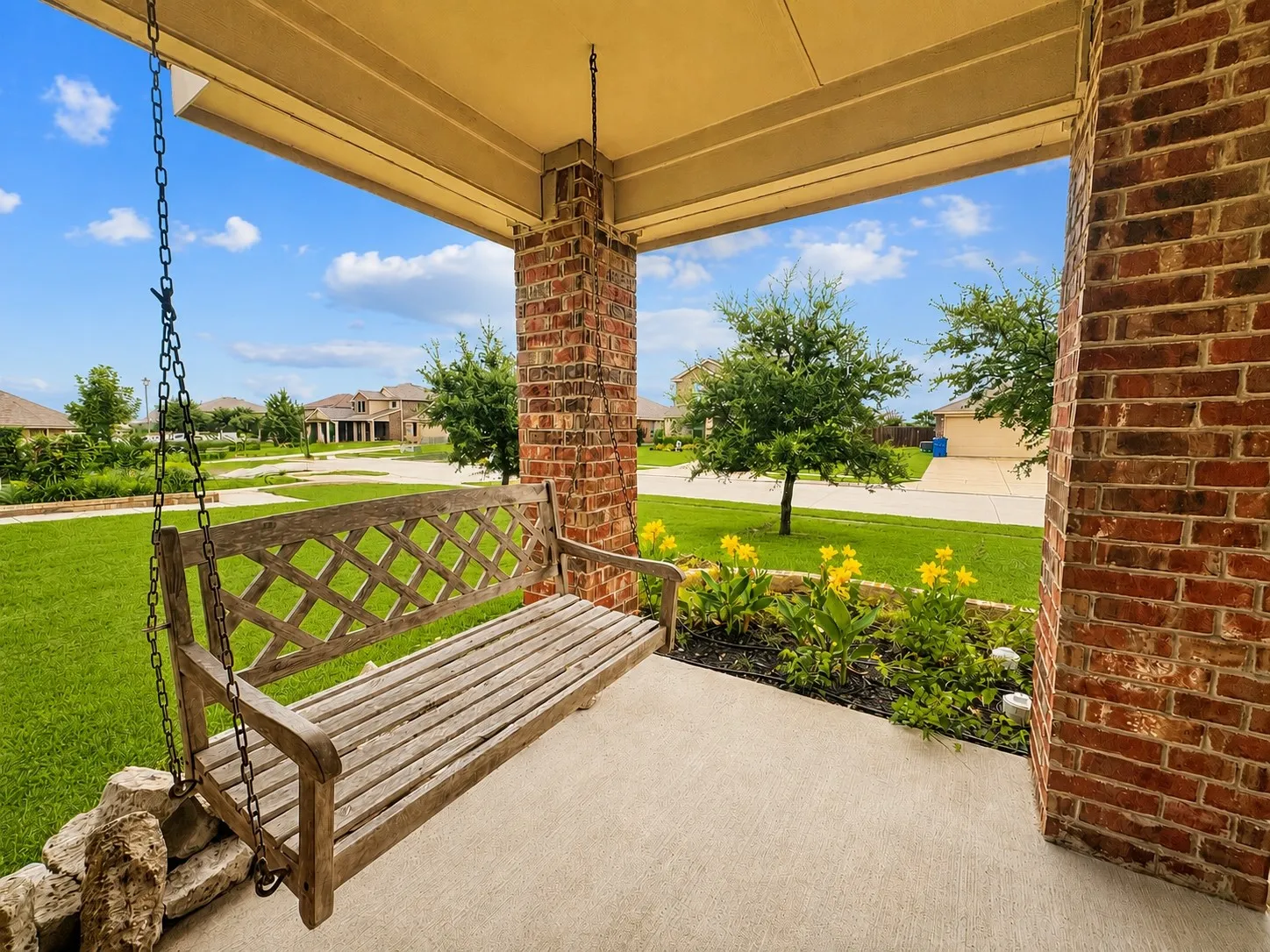Covered porch featuring a swing, brick columns, concrete flooring, and a landscaped garden bed with yellow flowering plants