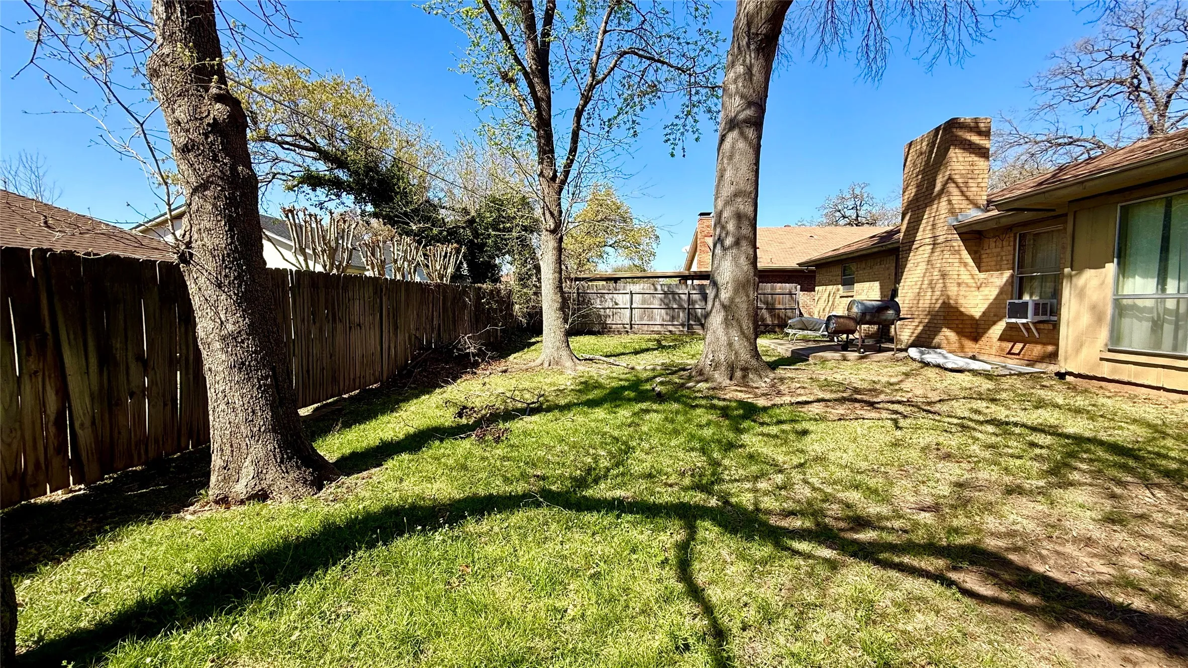 Spacious backyard with a wood privacy fence, mature trees, a brick chimney, and a brown shingle roof
