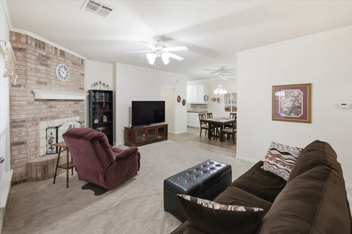 Spacious living area featuring a brick-clad fireplace with a white mantel, ceiling fan with integrated lighting, and neutral carpeting