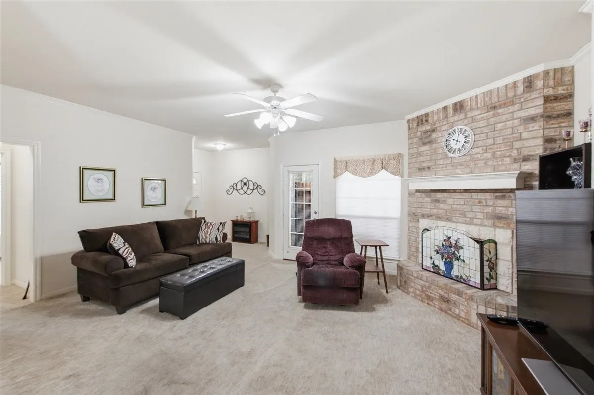 Living room featuring a brick fireplace with a decorative mantel, light-colored carpeting, a ceiling fan, and white walls