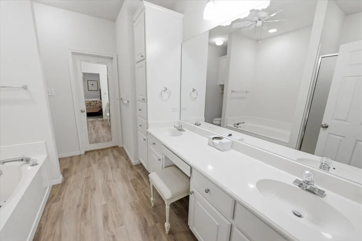 Primary bathroom featuring a double vanity with white cabinetry and integrated sinks, a built-in makeup vanity, a large wall-mounted mirror, wood-finish flooring, and a built-in bathtub and separate shower