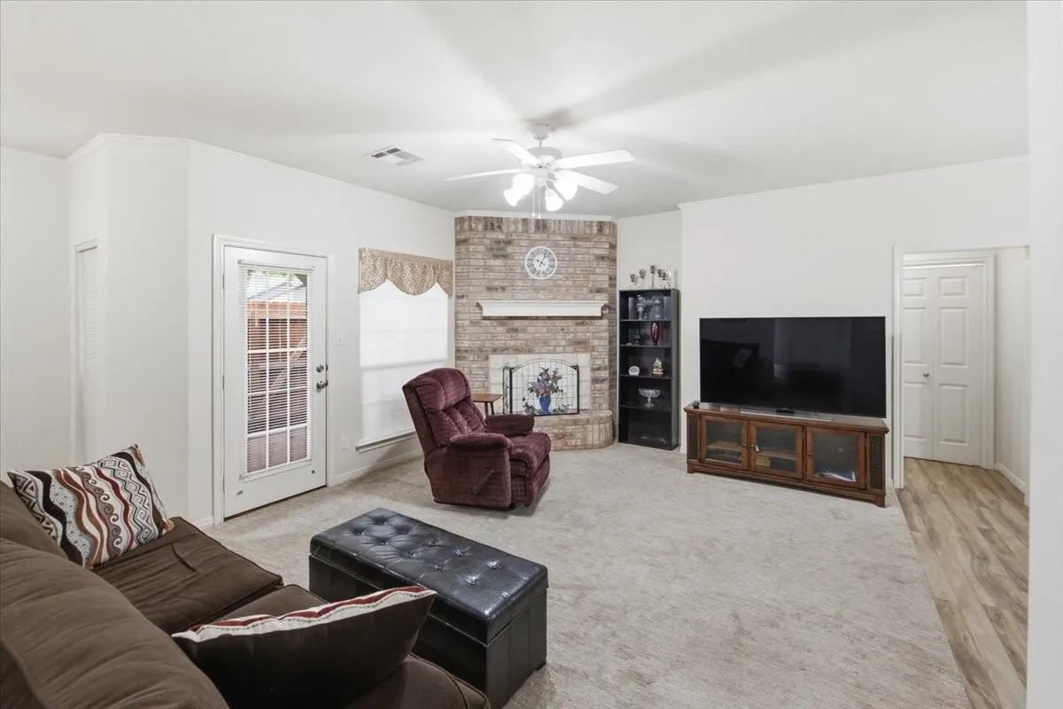 Spacious living area featuring light-colored carpeting, a corner brick fireplace, and a white ceiling fan