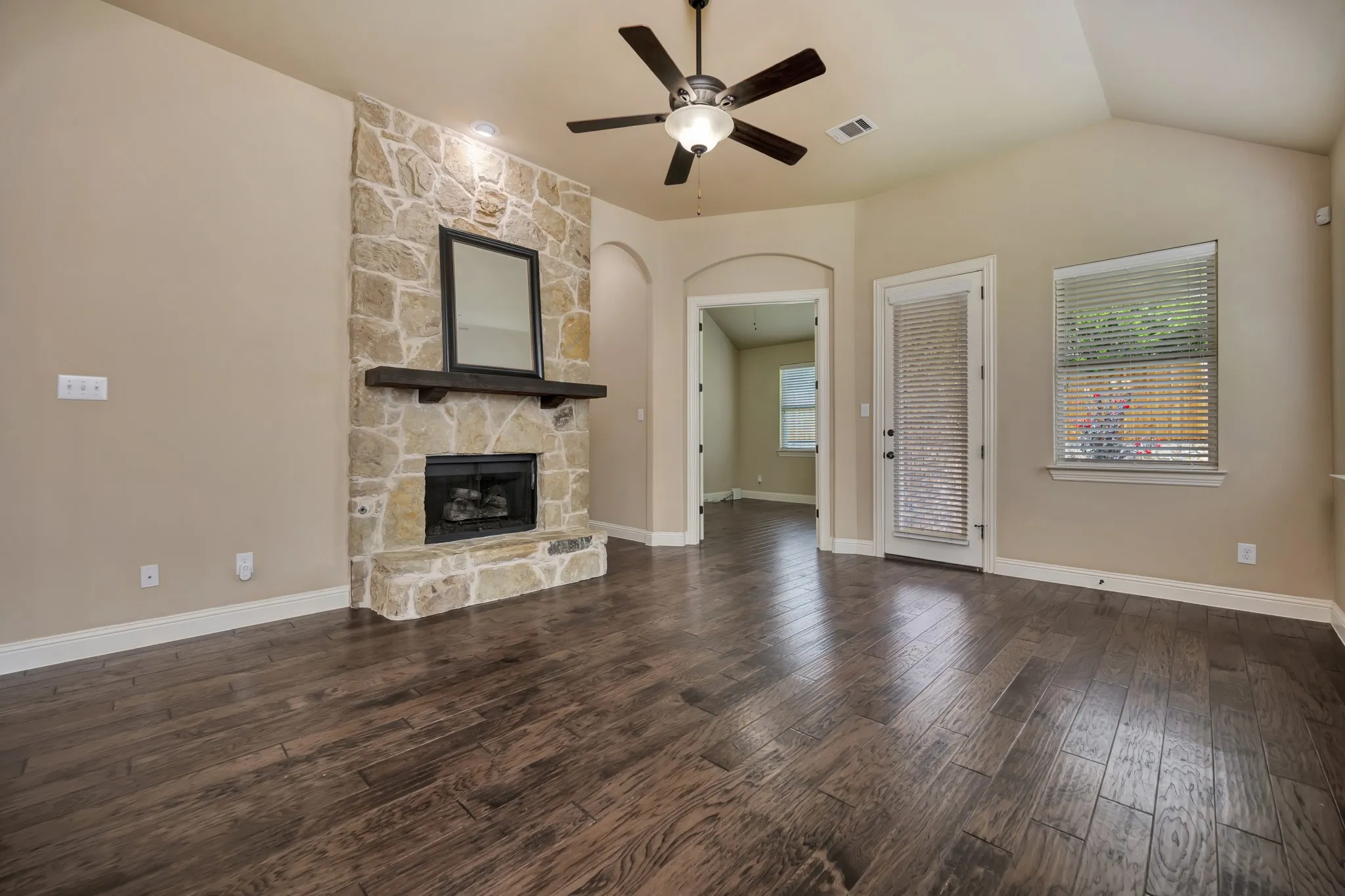 Spacious living area featuring a prominent stone fireplace with a wood mantel, dark wood-finish flooring, and a ceiling fan with integrated lighting