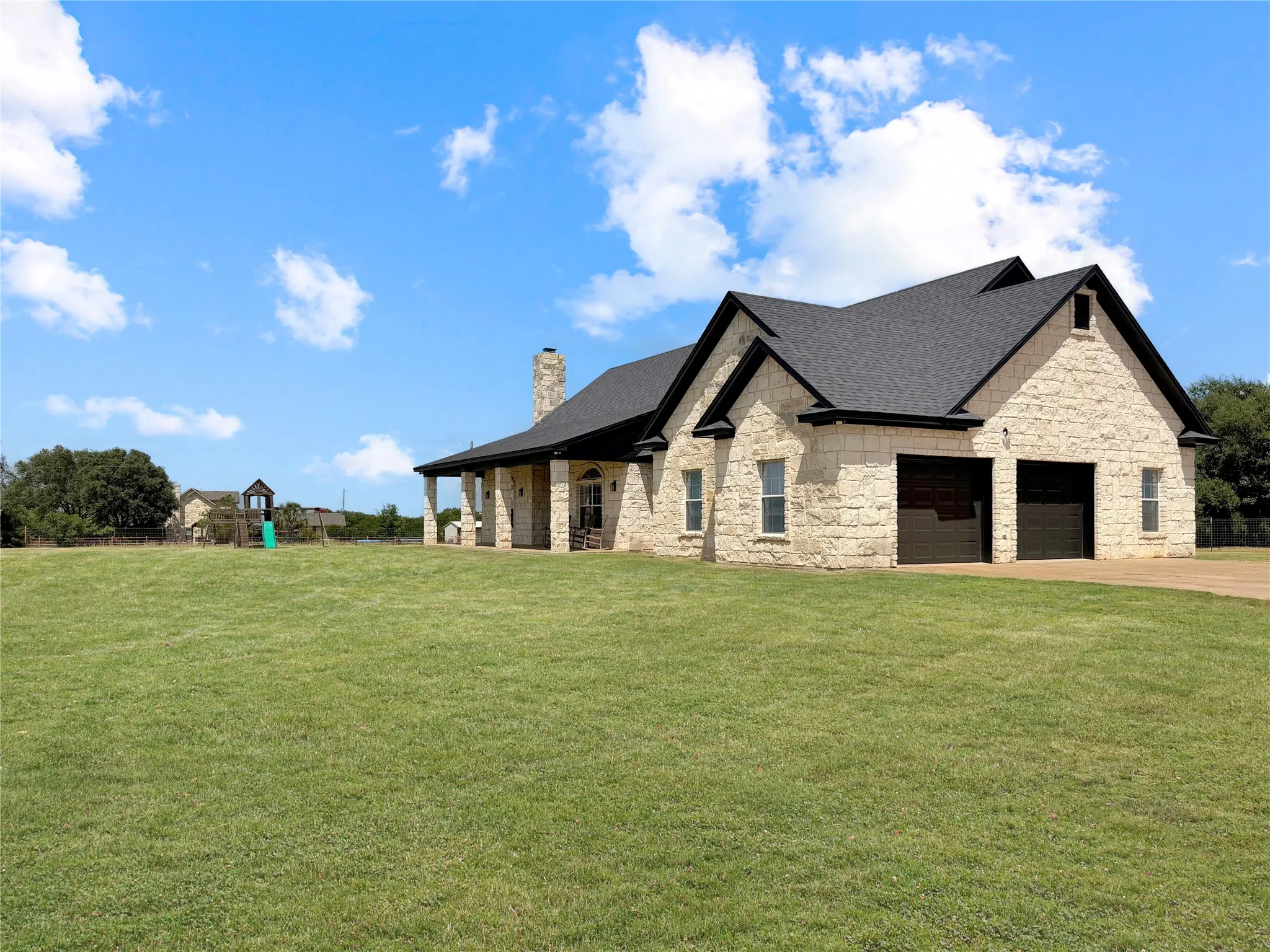 Stone exterior residence featuring a dark shingle roof, attached two-car garage with dark doors, a covered patio with columns, and a stone chimney