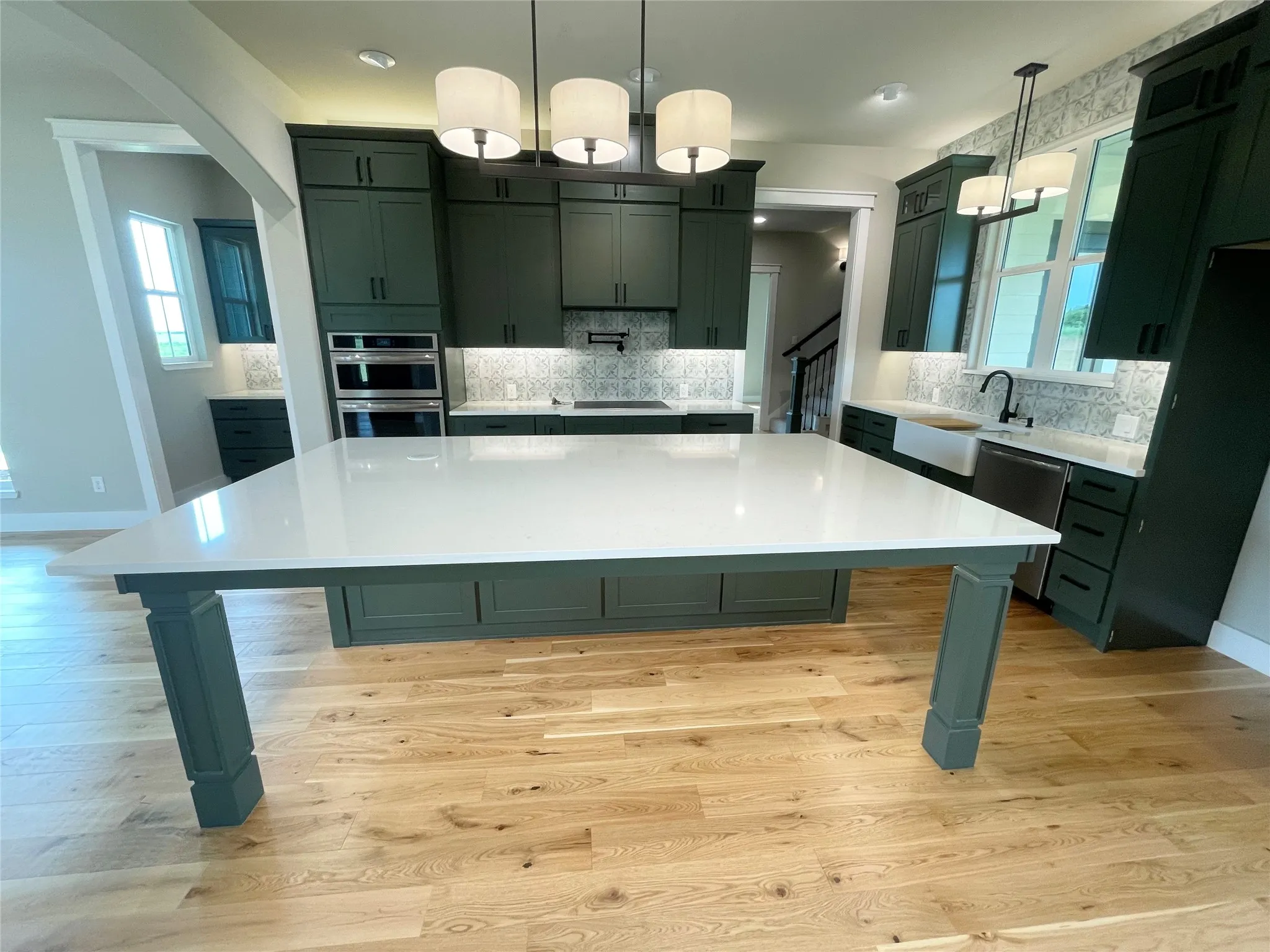 Spacious kitchen featuring an expansive central island with a light-toned countertop, deep green cabinetry, wood-finish flooring, a farmhouse sink, and integrated stainless steel appliances