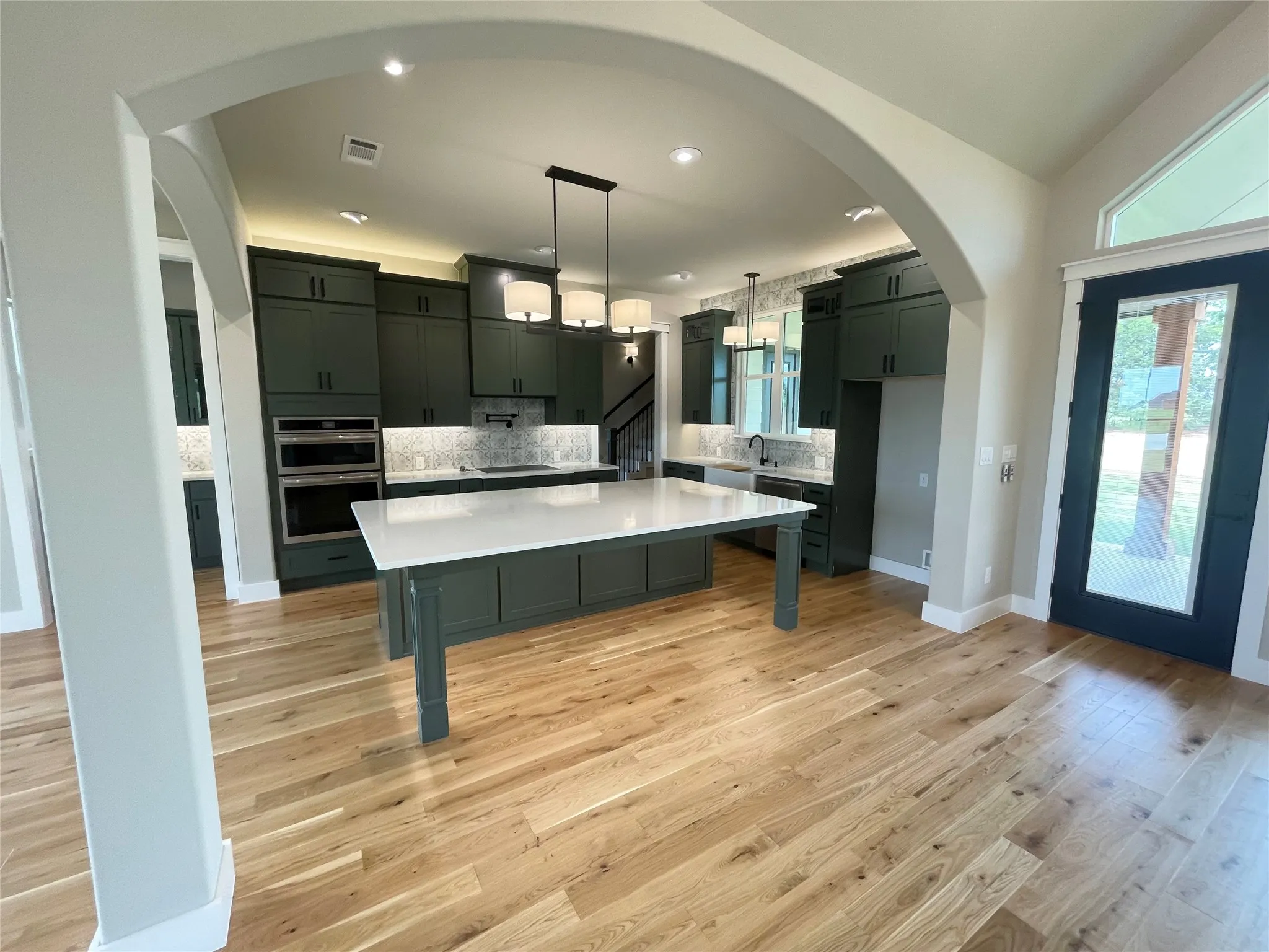 Open concept kitchen featuring dark green cabinetry, a central island with a white countertop, stainless steel double ovens, and light wood-finish flooring