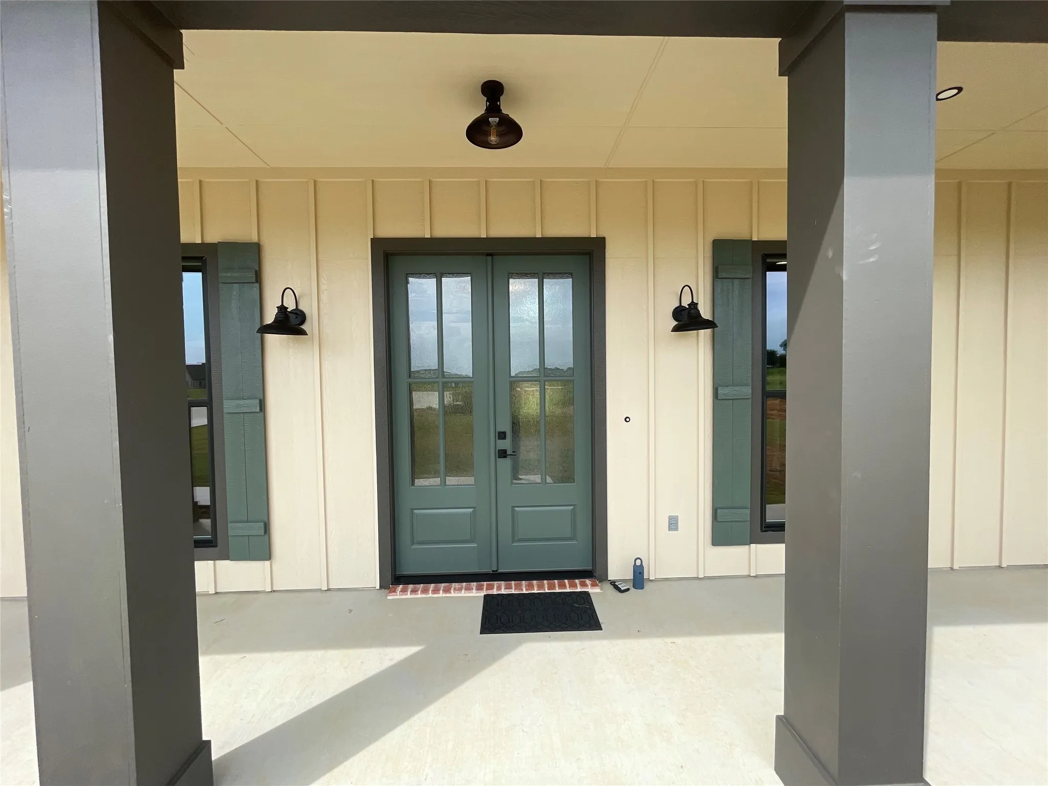 Covered front porch featuring board and batten siding, substantial support columns, double glass-panel entry doors, and wall-mounted exterior lighting