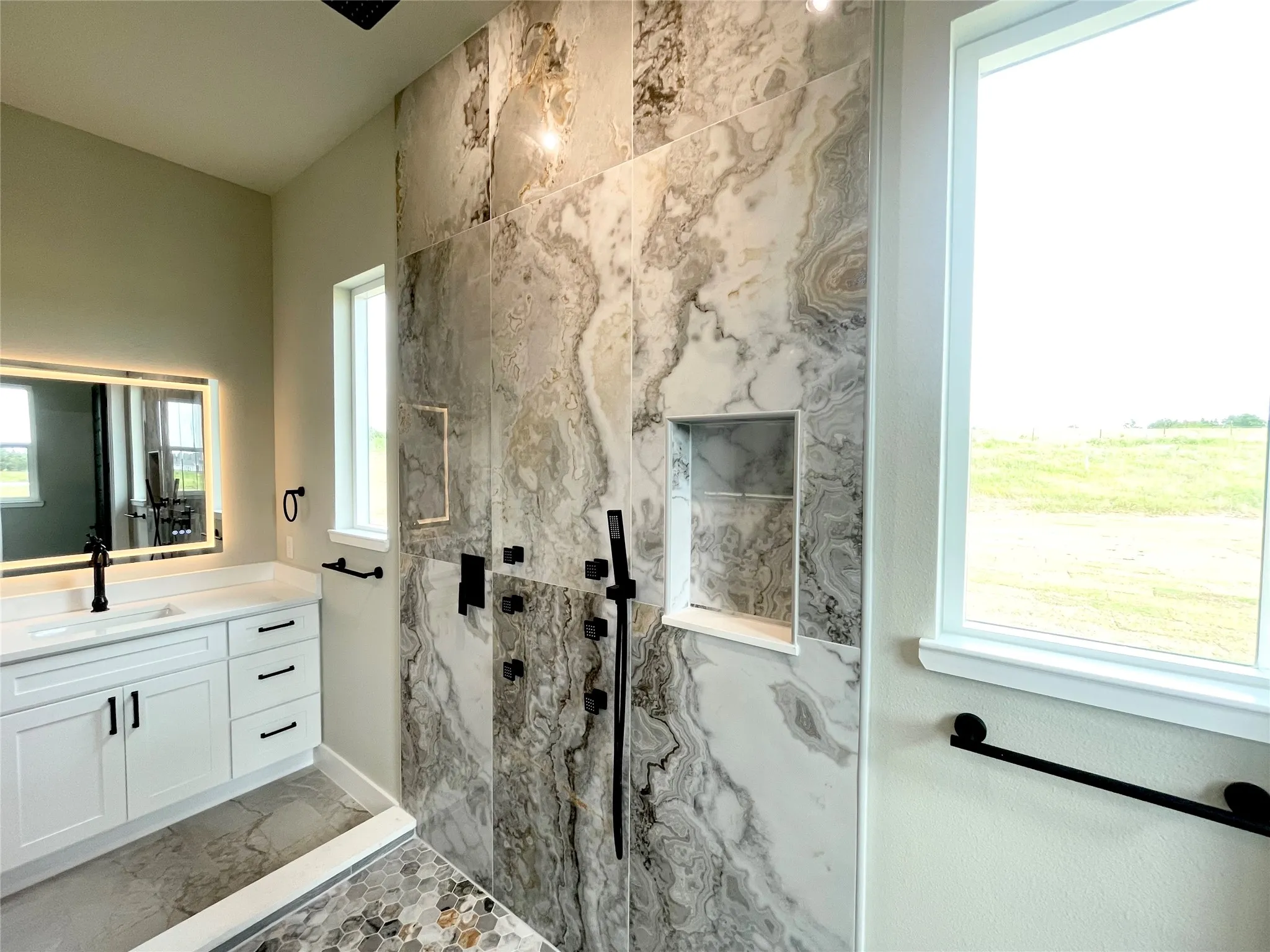 Walk-in shower with large format marble-look tile, mosaic floor, two recessed niches, and matte black fixtures