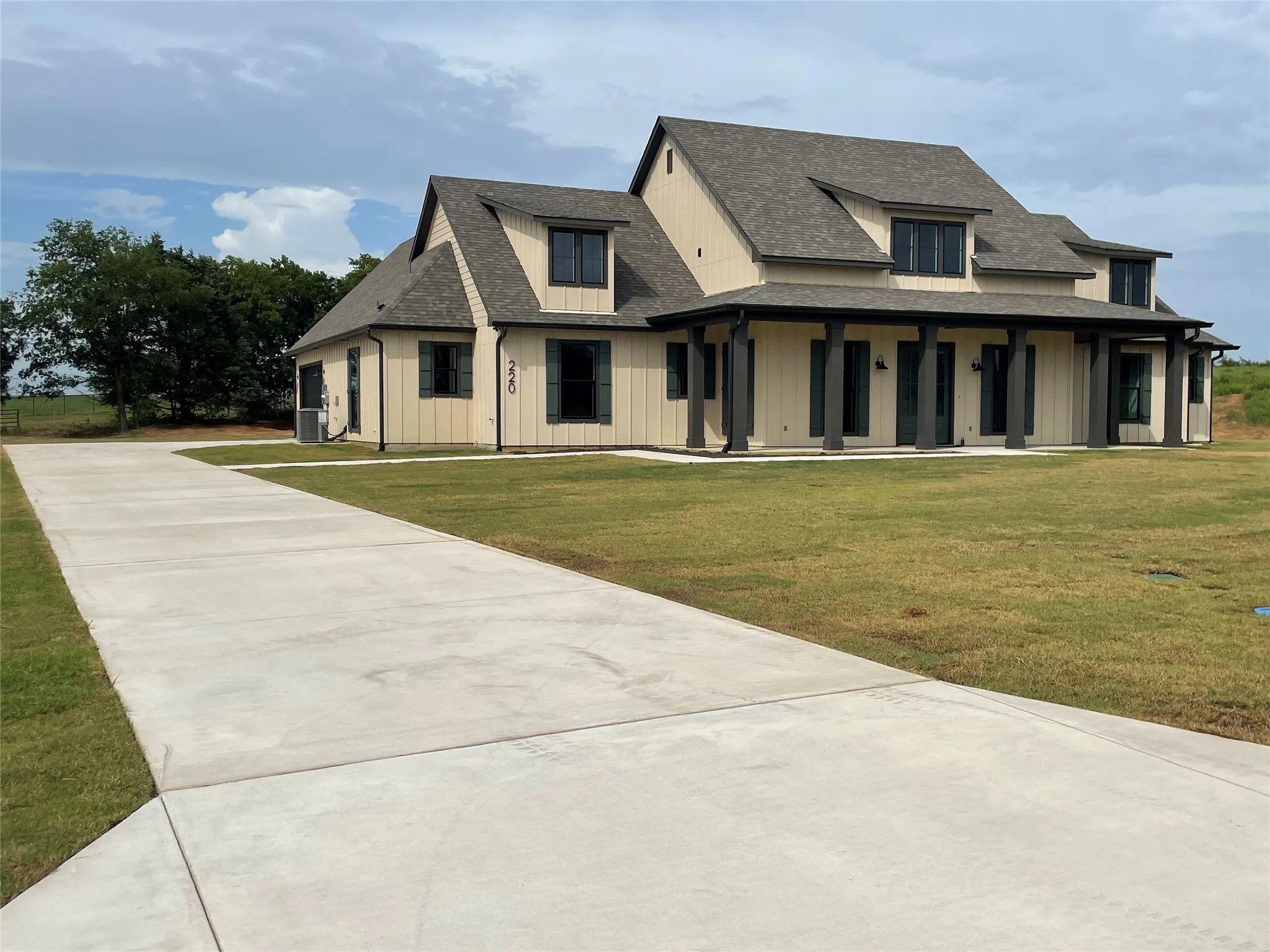 Contemporary farmhouse exterior featuring light-toned vertical siding, dark window trim, a covered front porch with substantial columns, an asphalt shingle roof, and a concrete driveway