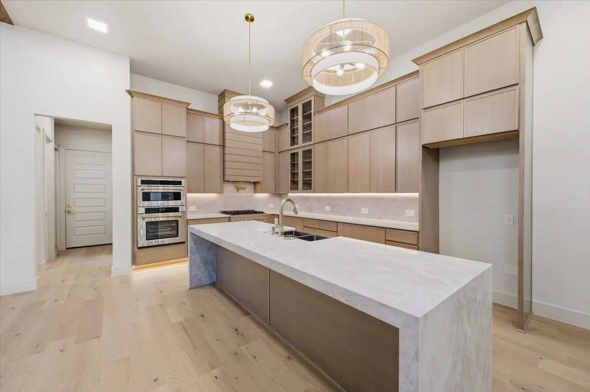 Gourmet kitchen featuring a waterfall island with a stone countertop, light wood-finish cabinetry, integrated double ovens, and wood-finish flooring