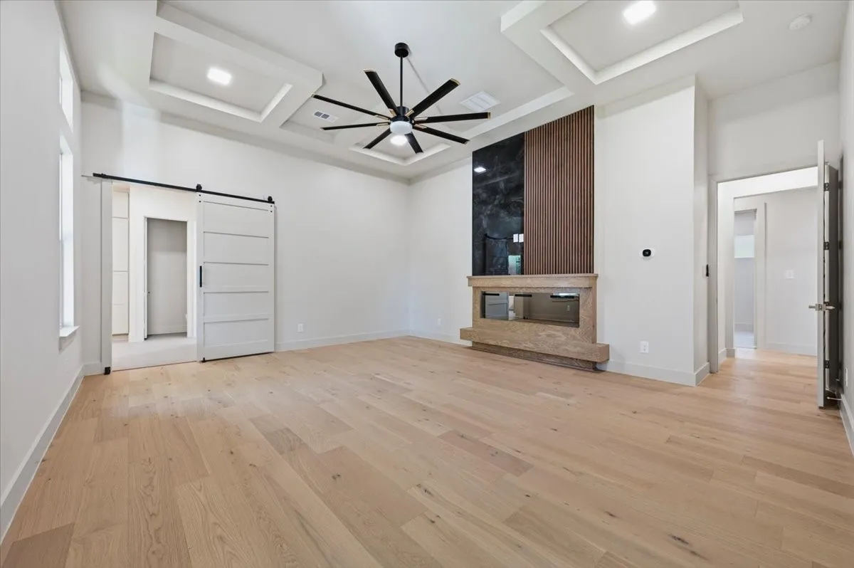 Spacious room featuring light wood-finish flooring, a contemporary fireplace with a dark stone and wood-slat accent wall, and a multi-tiered tray ceiling with recessed lighting and a modern ceiling fan