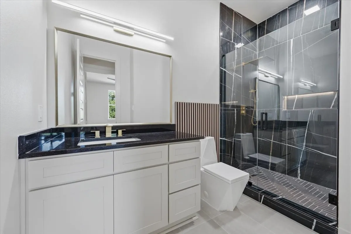 Modern bathroom featuring a frameless glass shower enclosure with dark veined tile, a white vanity with dark countertop and undermount sink, and light-toned floor tiling