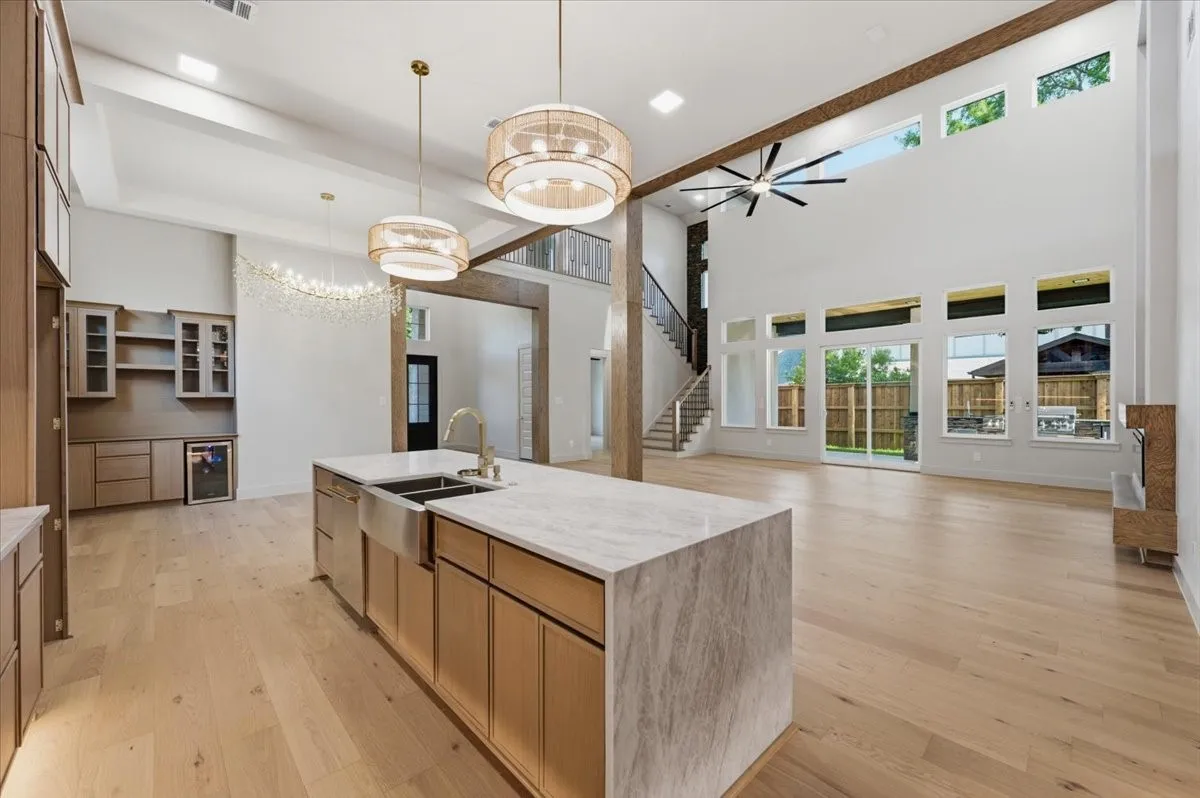 Spacious open-concept interior featuring light wood-finish flooring, a kitchen island with a waterfall countertop and stainless steel sink, a beverage bar with glass-front cabinetry, a two-story living area with expansive windows, and a multi-level staircase with wood treads
