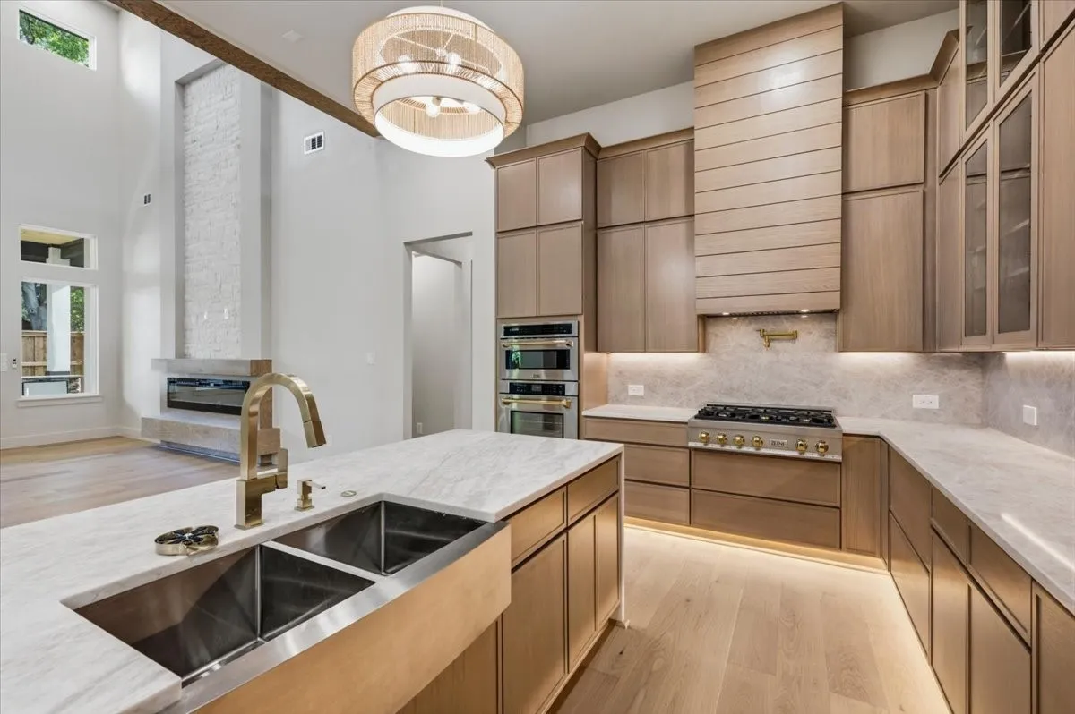 Gourmet kitchen featuring a large island with an undermount double basin sink and brass faucet, light wood-finish cabinetry, built-in stainless steel appliances, and under-cabinet lighting