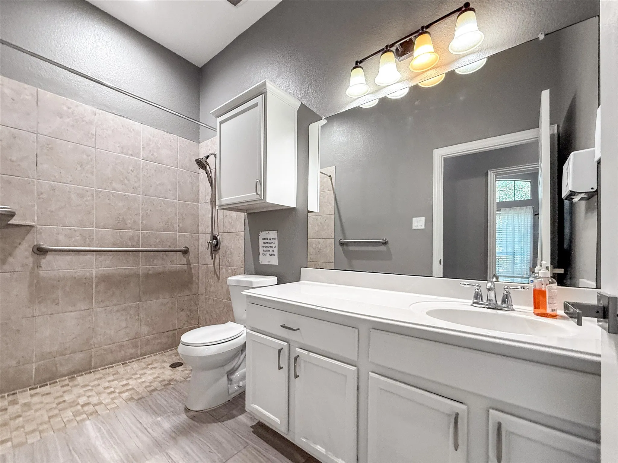 Bathroom featuring a vanity with a white countertop and double white cabinetry, a large wall-mounted mirror, a four-light fixture, and a tiled shower with grab bars and a handheld showerhead