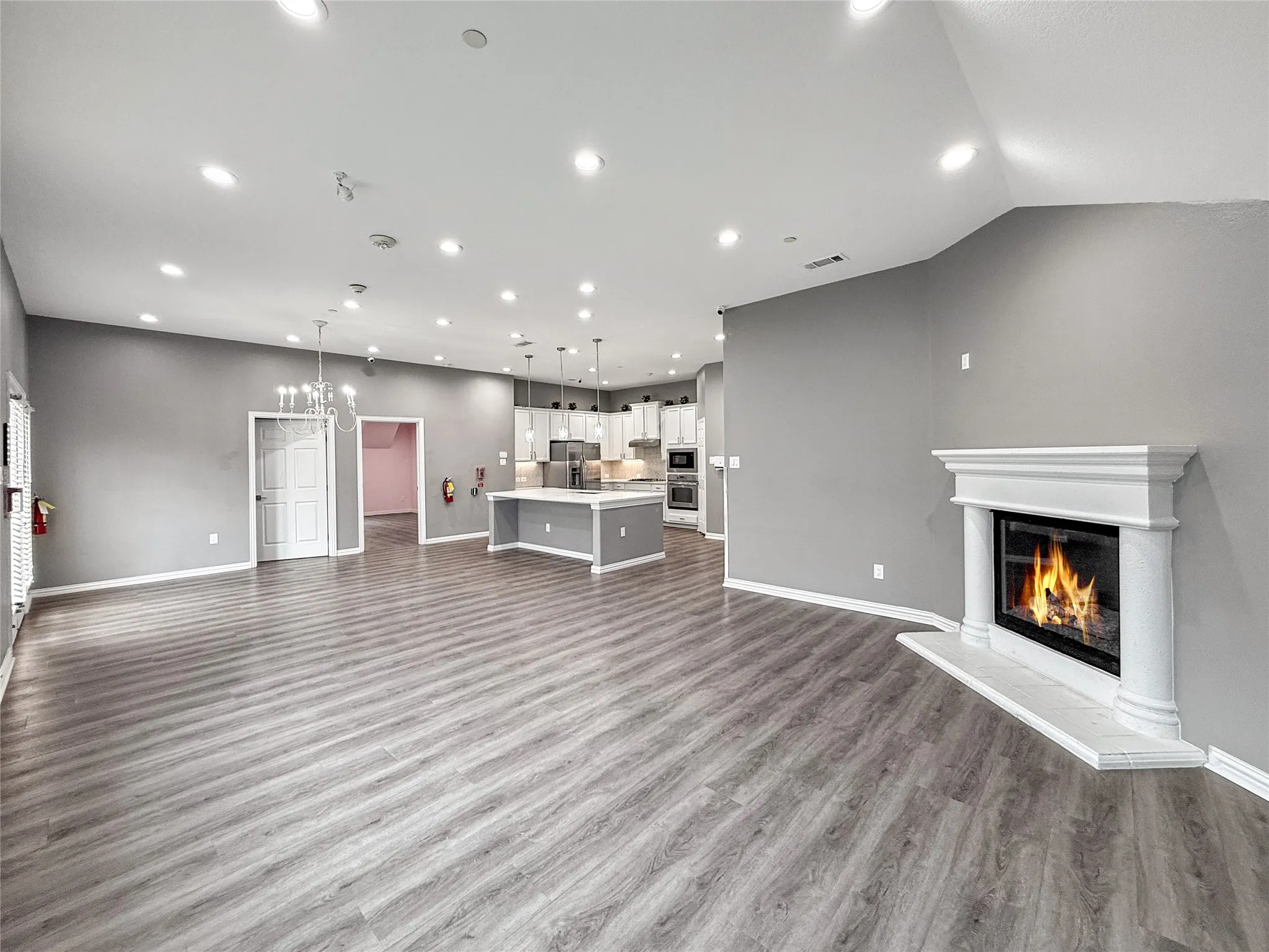 Spacious open-concept interior featuring light wood-finish flooring, a white fireplace with an ornate mantel, and recessed ceiling lighting