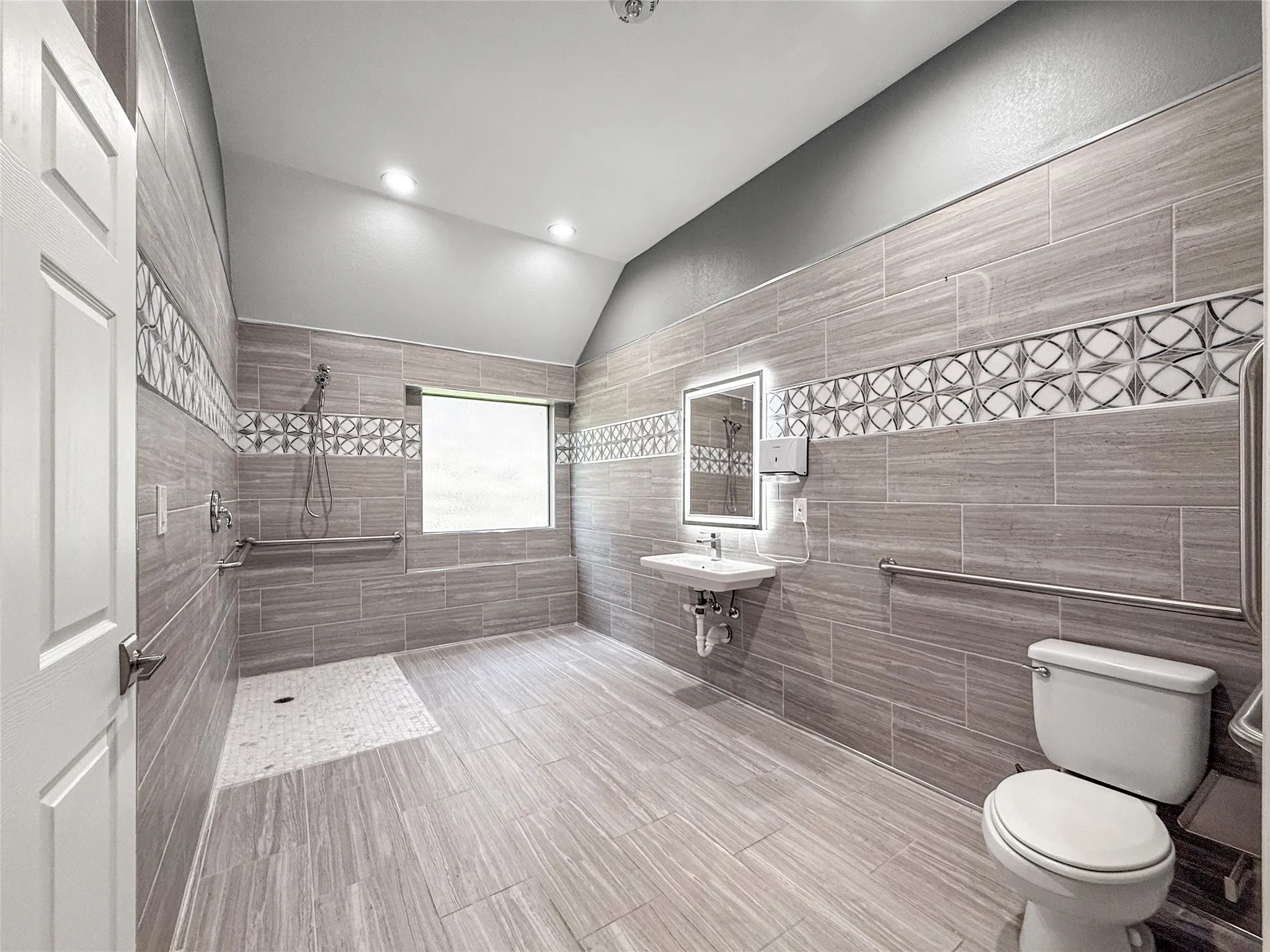 Spacious full bathroom featuring floor-to-ceiling tile, a walk-in shower with grab bars, a floating sink vanity, an illuminated mirror, and a window for natural light