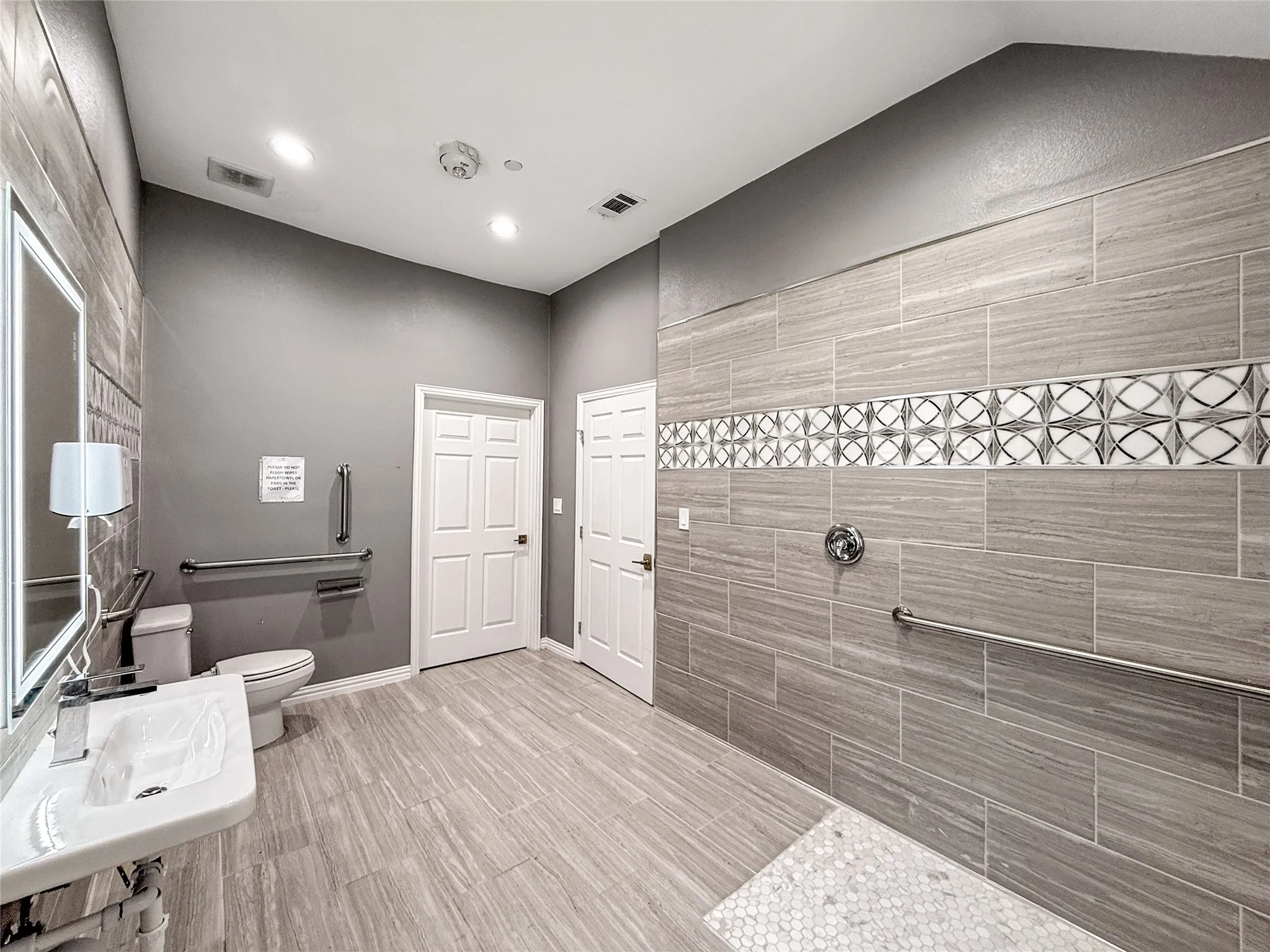 Spacious bathroom featuring large format wood-look tile flooring, a wall-mounted sink, and a walk-in shower with decorative mosaic tile accent