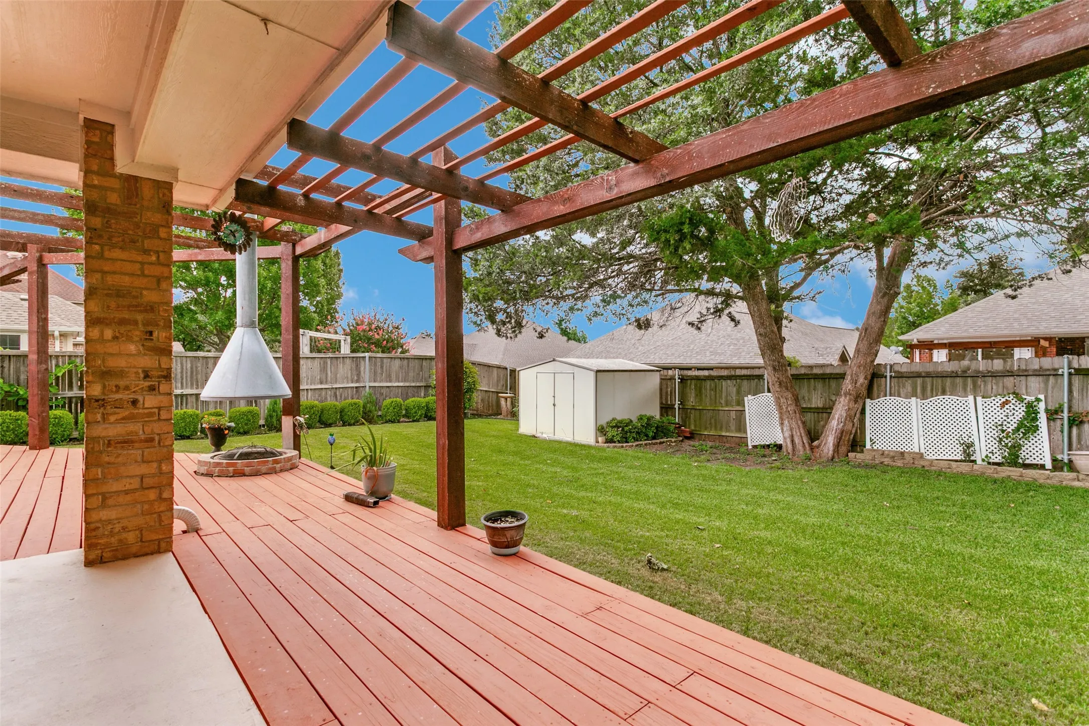 Expansive backyard featuring a wood-finish deck with pergola, a built-in outdoor fireplace, a manicured lawn, and mature trees