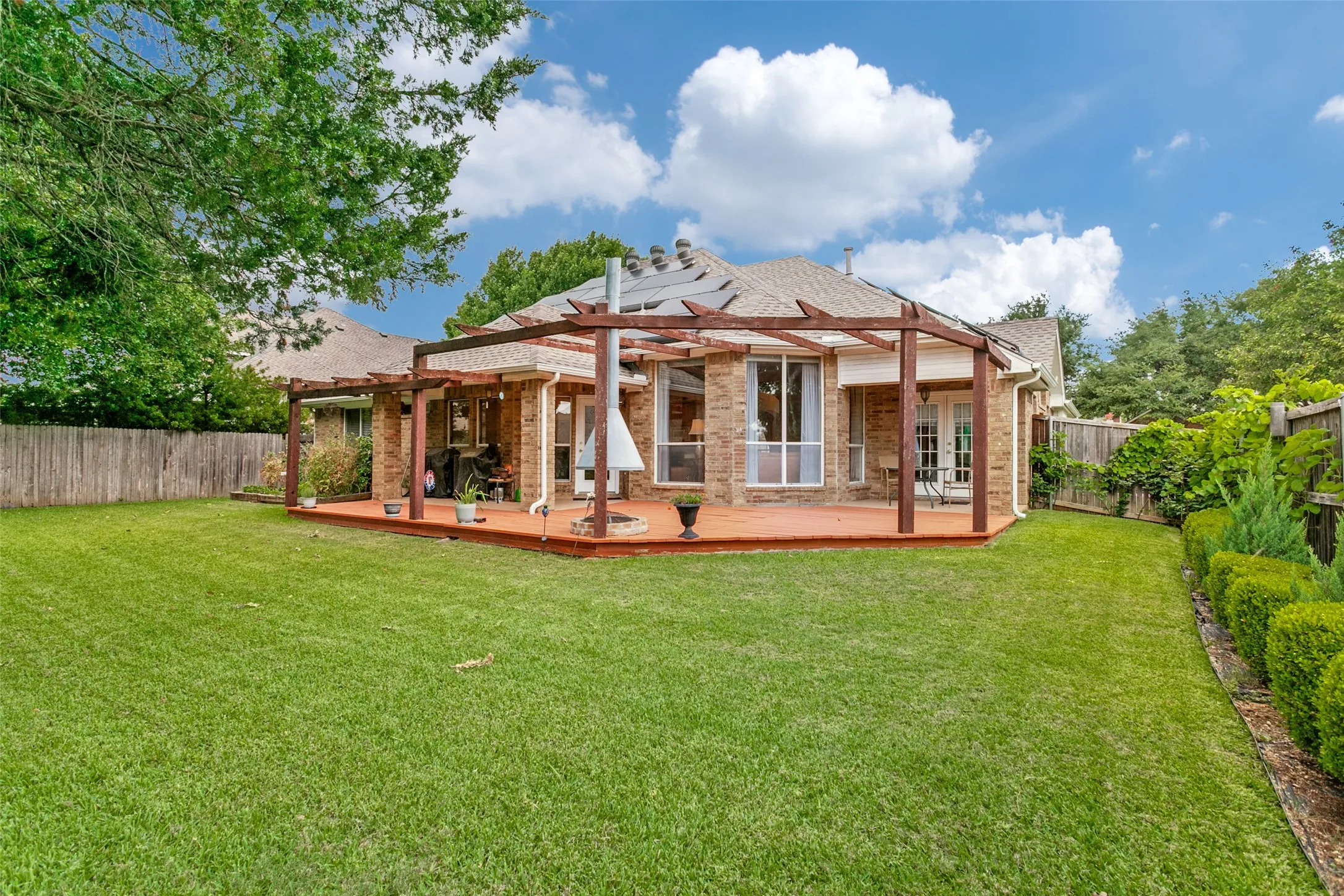 Expansive backyard featuring a large wooden deck with a pergola, brick exterior, multiple windows, and a privacy fence
