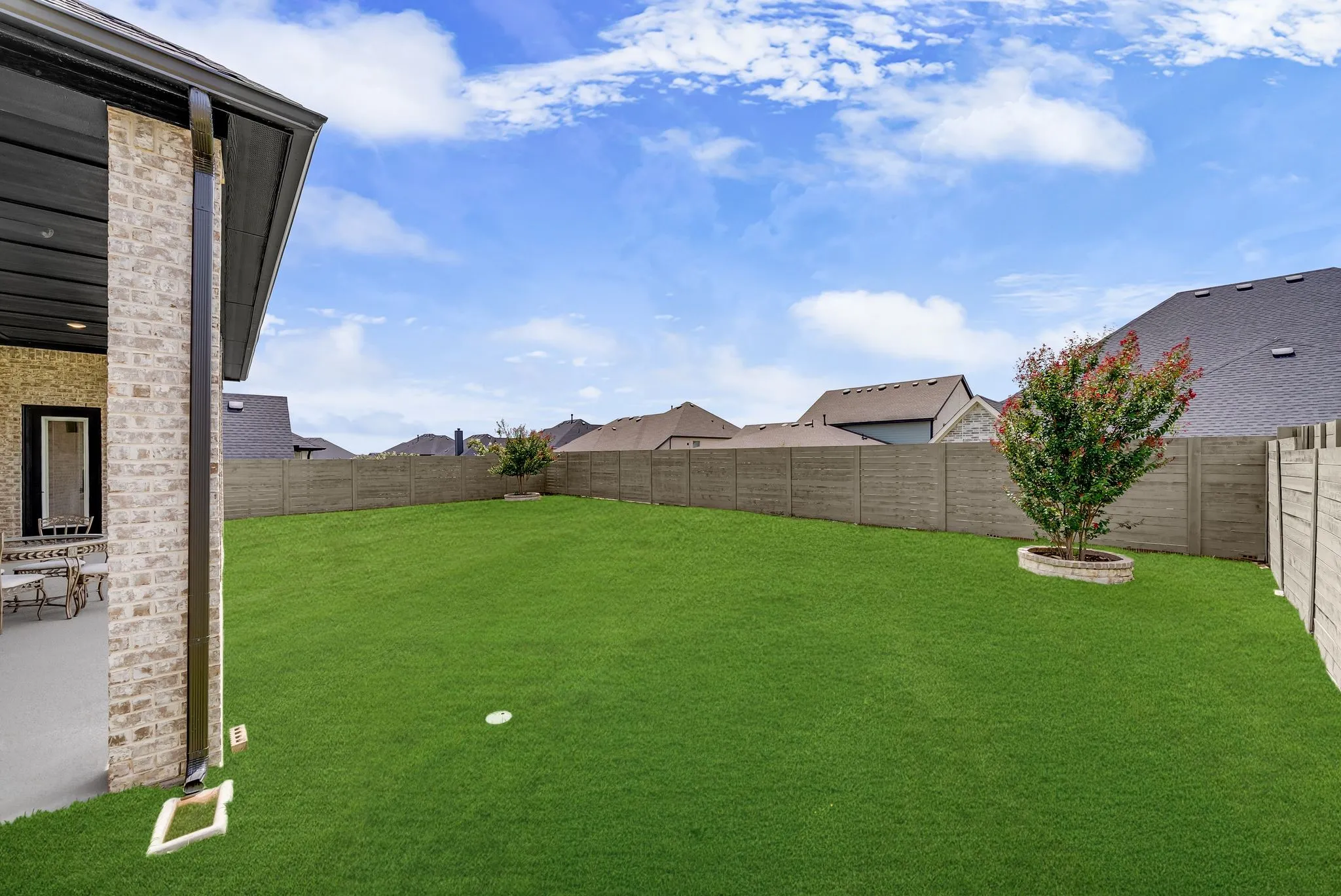 Expansive green lawn with a perimeter fence, offering a covered patio with brick exterior and a small tree with stone-edged planting