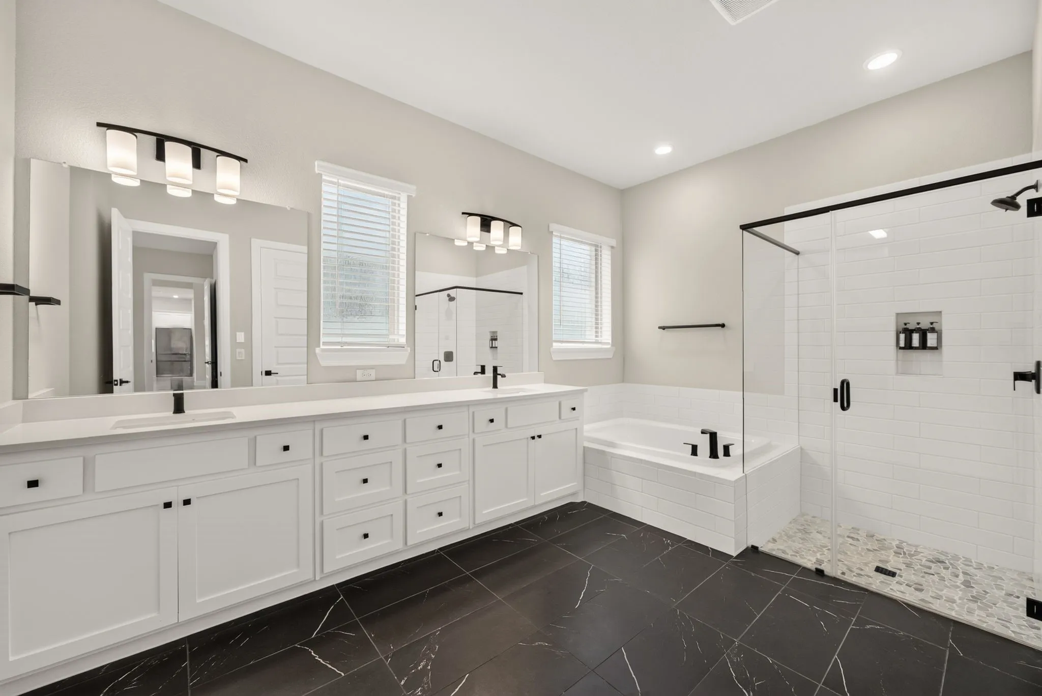 Spacious bathroom featuring a dual vanity with white cabinetry and countertops, a built-in soaking tub, a glass-enclosed shower with subway tile and pebble flooring, and large format dark tile flooring