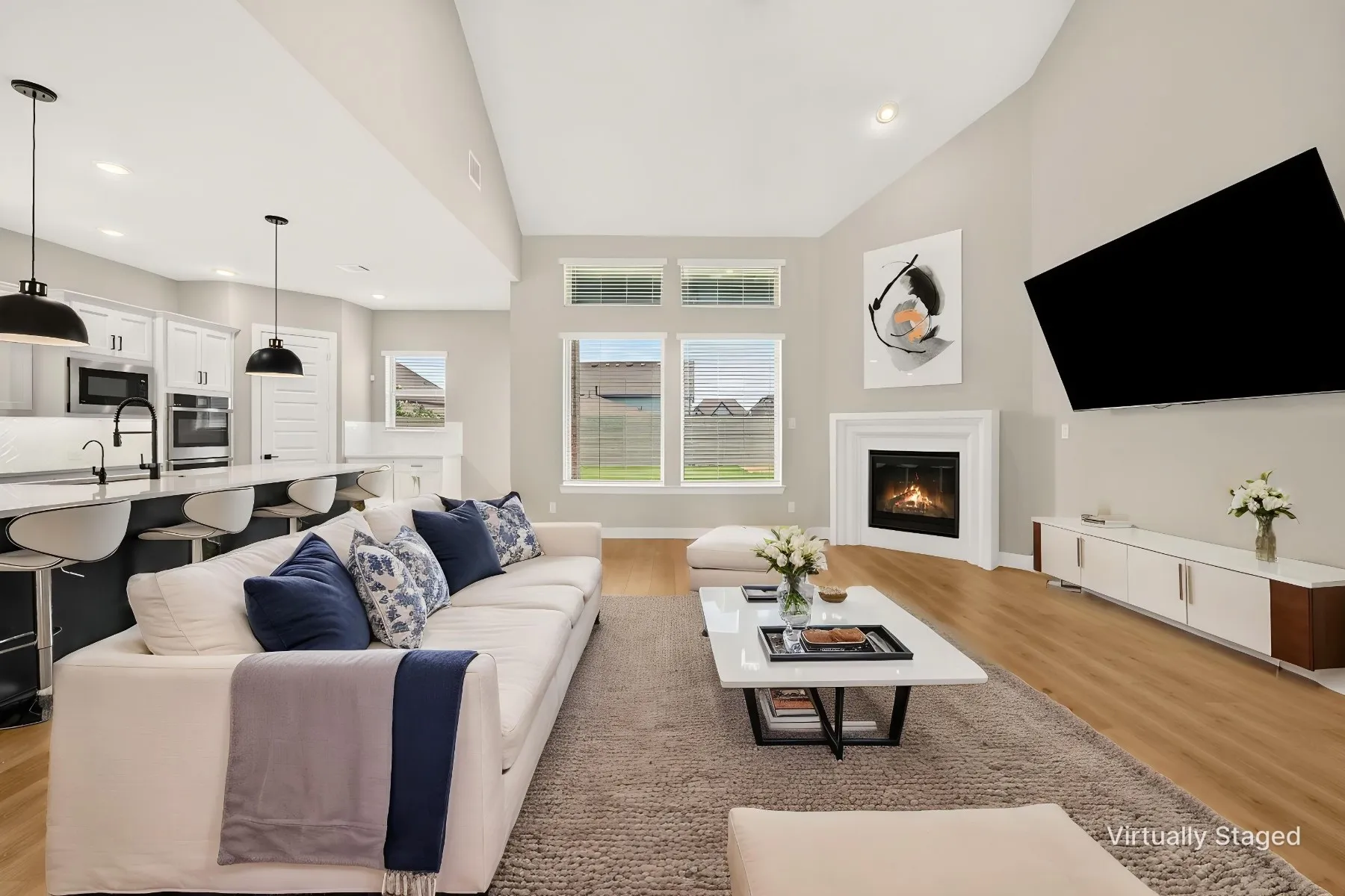Open-concept living area featuring high vaulted ceilings, wood-finish flooring, a contemporary fireplace with white mantel, a kitchen island with seating, and ample natural light from multiple windows