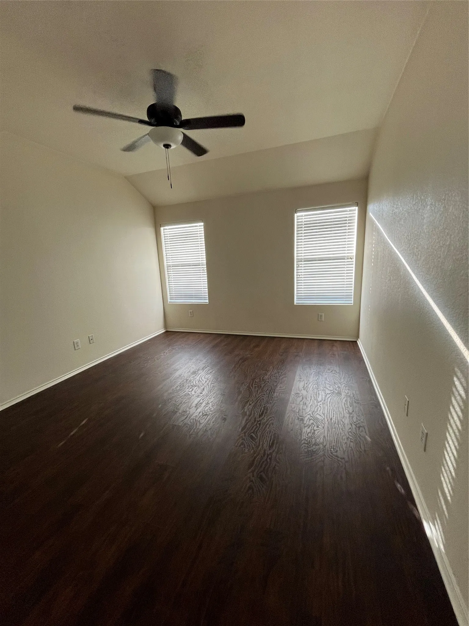 Spacious room featuring wood-finish flooring, two windows with blinds, a ceiling fan with light fixture, and neutral wall tones