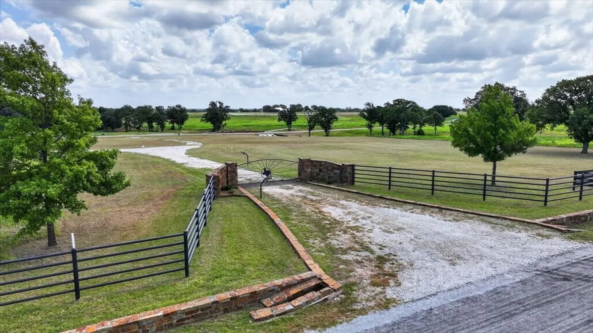 Expansive property featuring a gravel driveway, stone and wrought iron entry gate, black rail fencing, mature trees, and a vast grassy landscape