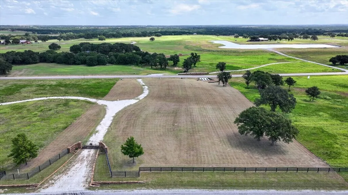 Expansive land parcel featuring a gravel driveway entrance with stone columns, partial fencing, mature trees, and a distant water feature