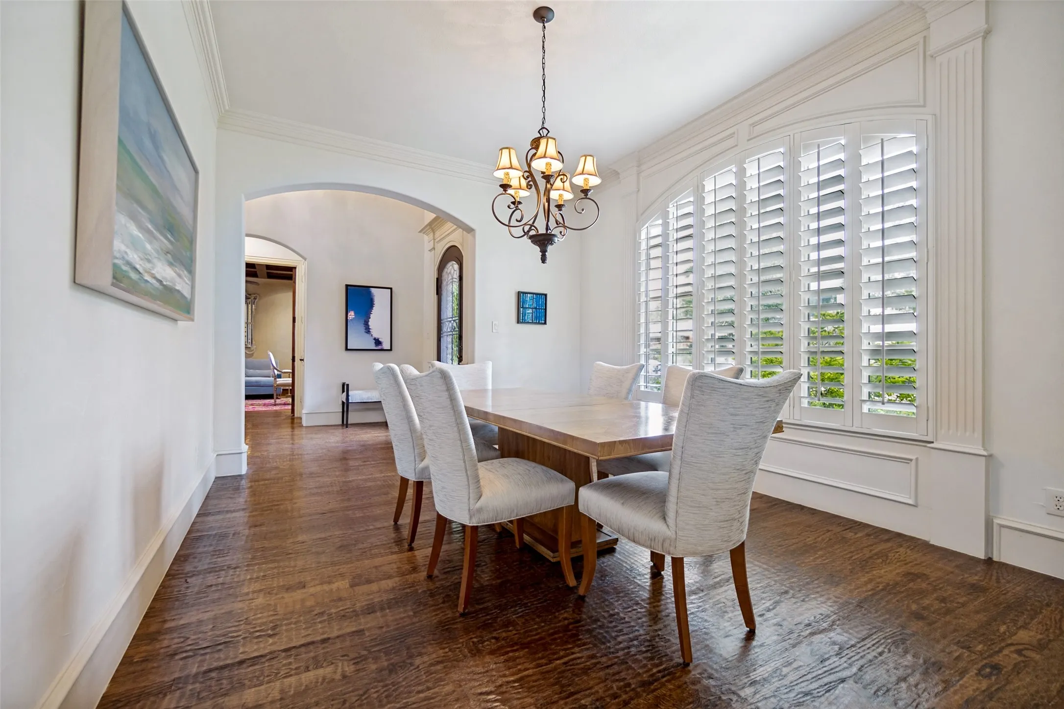 Formal dining area featuring rich wood-finish flooring, an arched window with plantation shutters, crown molding, and an elegant chandelier