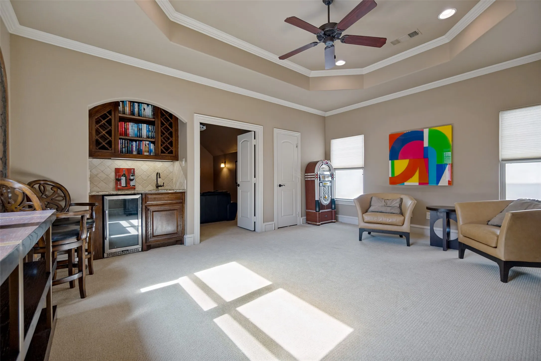 Spacious room with a tray ceiling and ceiling fan, featuring a built-in wet bar with wood cabinetry, integrated beverage refrigerator, and tile backsplash