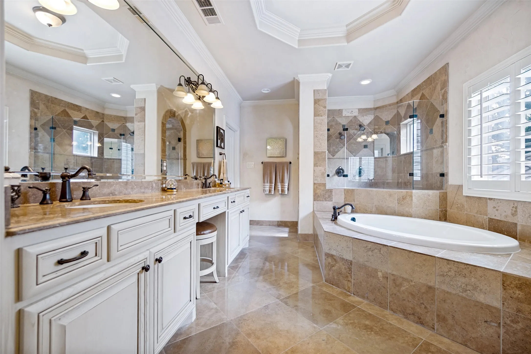 Expansive bathroom featuring a built-in soaking tub, a tiled walk-in shower with glass enclosure, dual vanity sinks with dark bronze fixtures, a vanity seating area, and plantation shutters