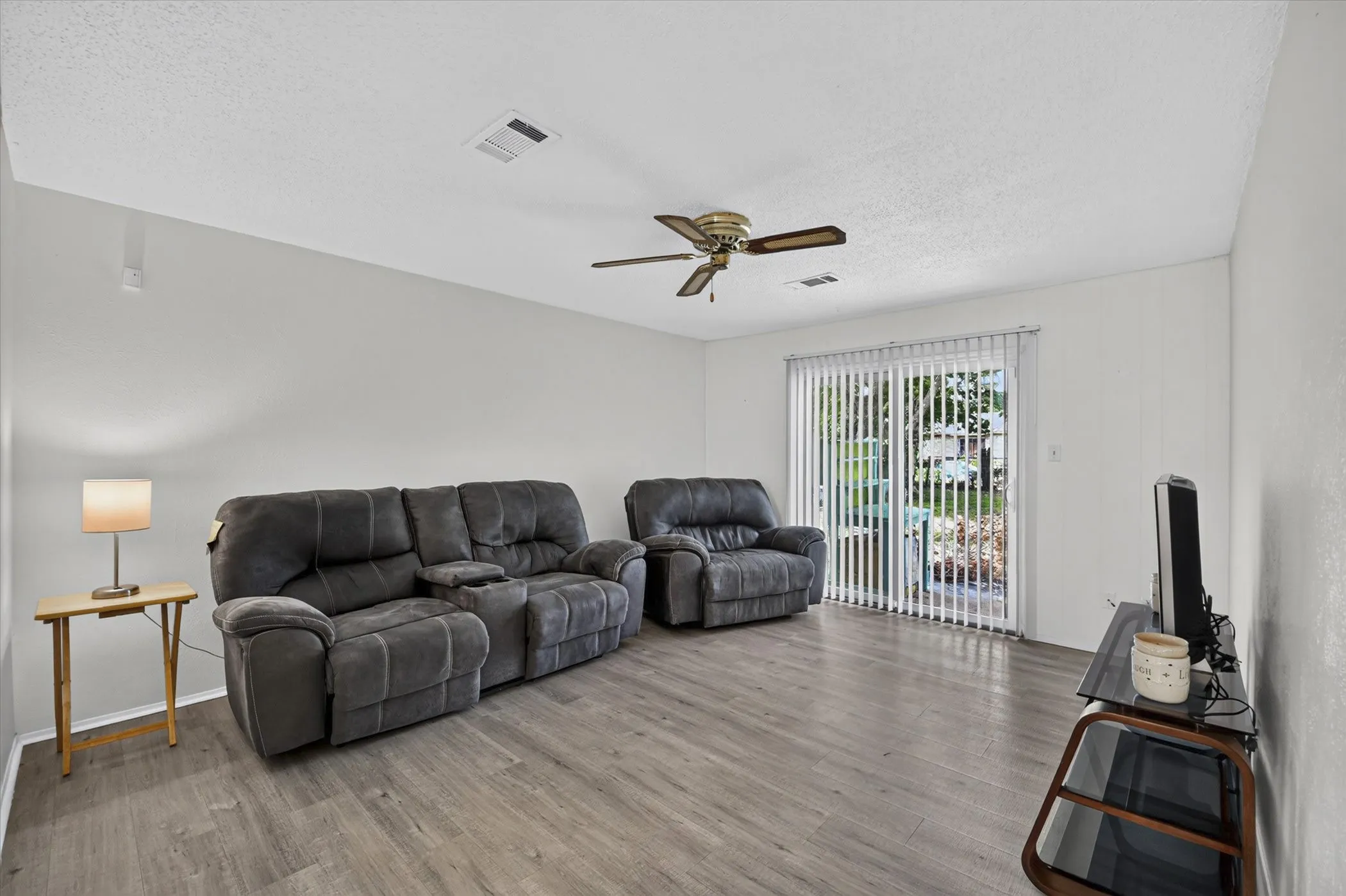 Spacious living area featuring wood-finish flooring, a ceiling fan, and a sliding glass door with vertical blinds providing exterior access