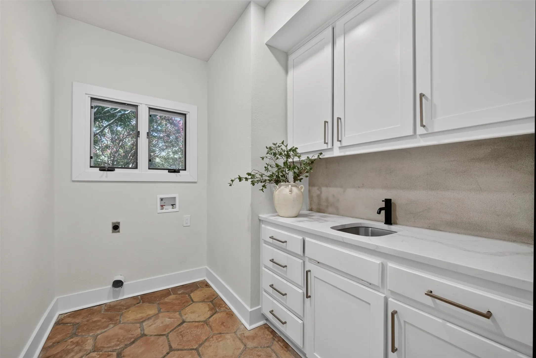 Laundry area featuring white cabinetry, a countertop with an undermount sink and matte black faucet, hexagonal terracotta flooring, and a window providing natural light