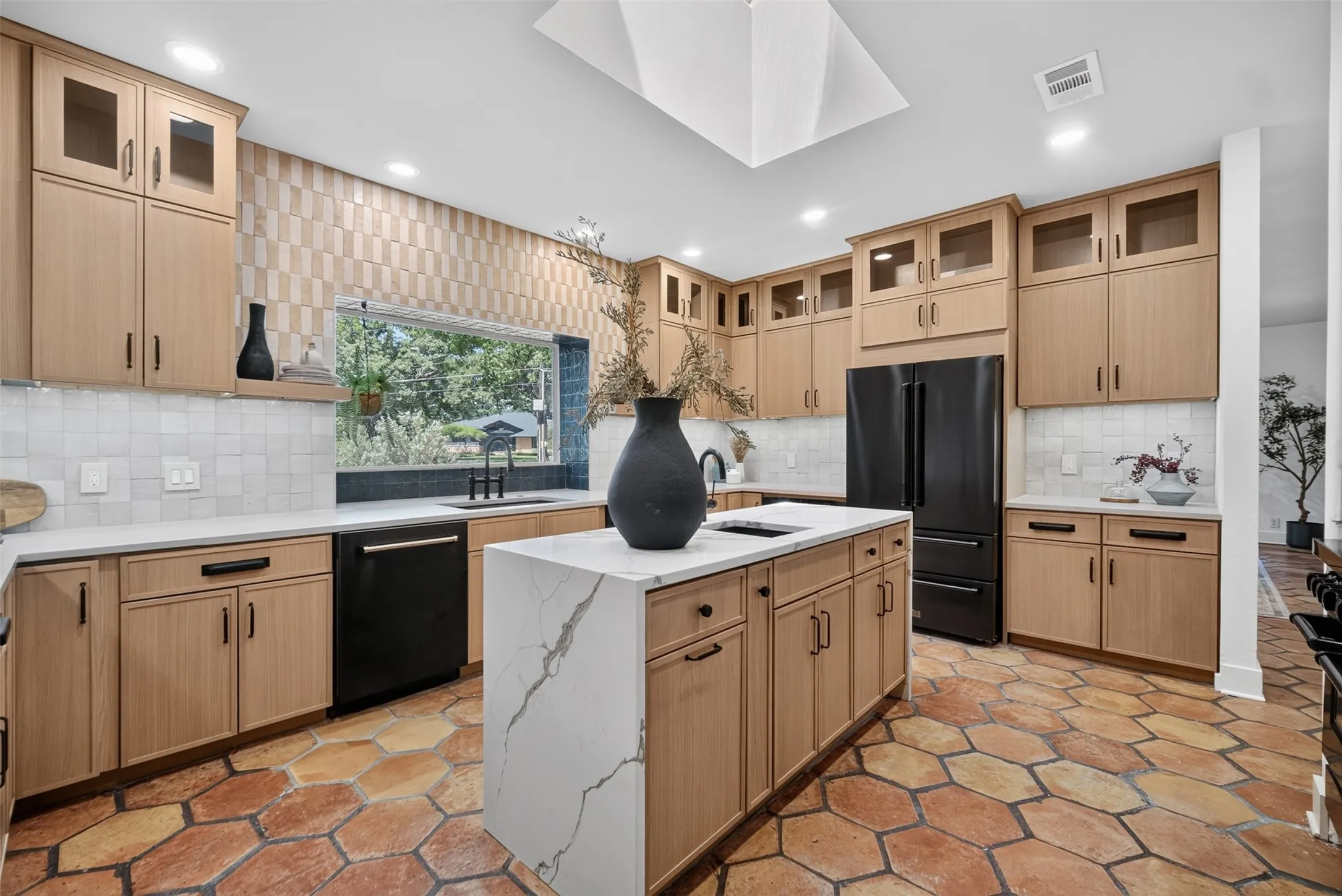 Spacious kitchen featuring an expansive central island with a waterfall countertop, abundant natural wood-finish cabinetry, a large skylight, hexagon tile flooring, and integrated black appliances