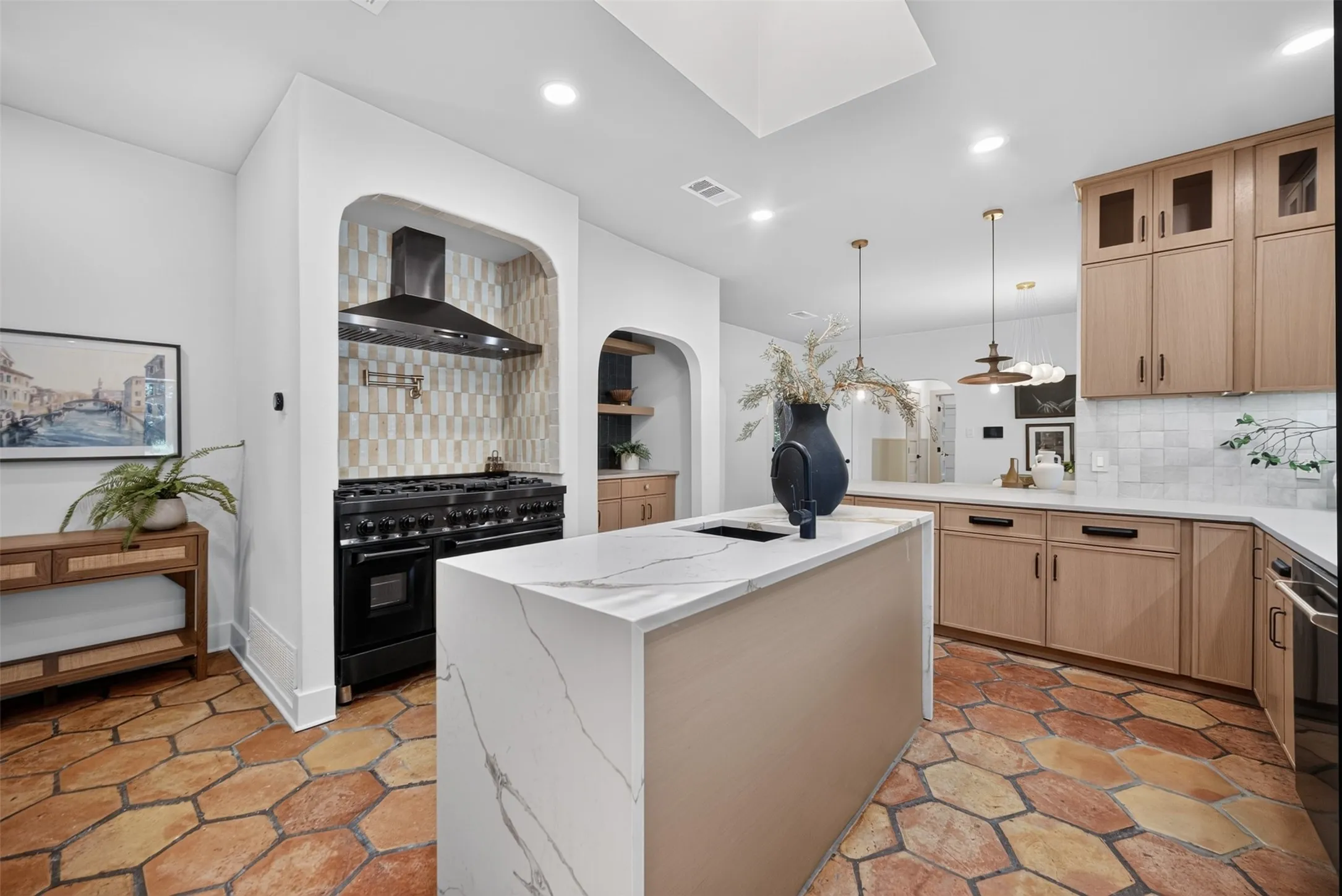 Kitchen featuring hexagonal terracotta-style flooring, a large island with a waterfall countertop and integrated sink, custom light wood-finish cabinetry, white tile backsplash, and a professional-grade black range with an arched alcove and patterned tile surround
