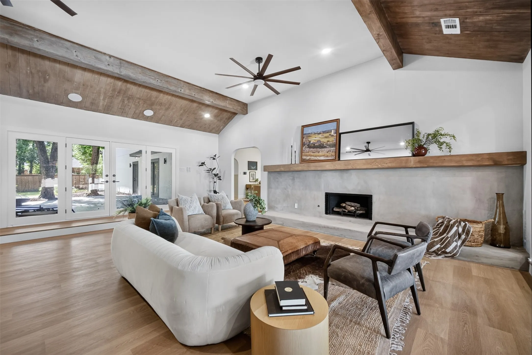 Spacious living area featuring a vaulted ceiling with exposed wood beams, a concrete-finish fireplace with a substantial wood mantel, and light wood-finish flooring