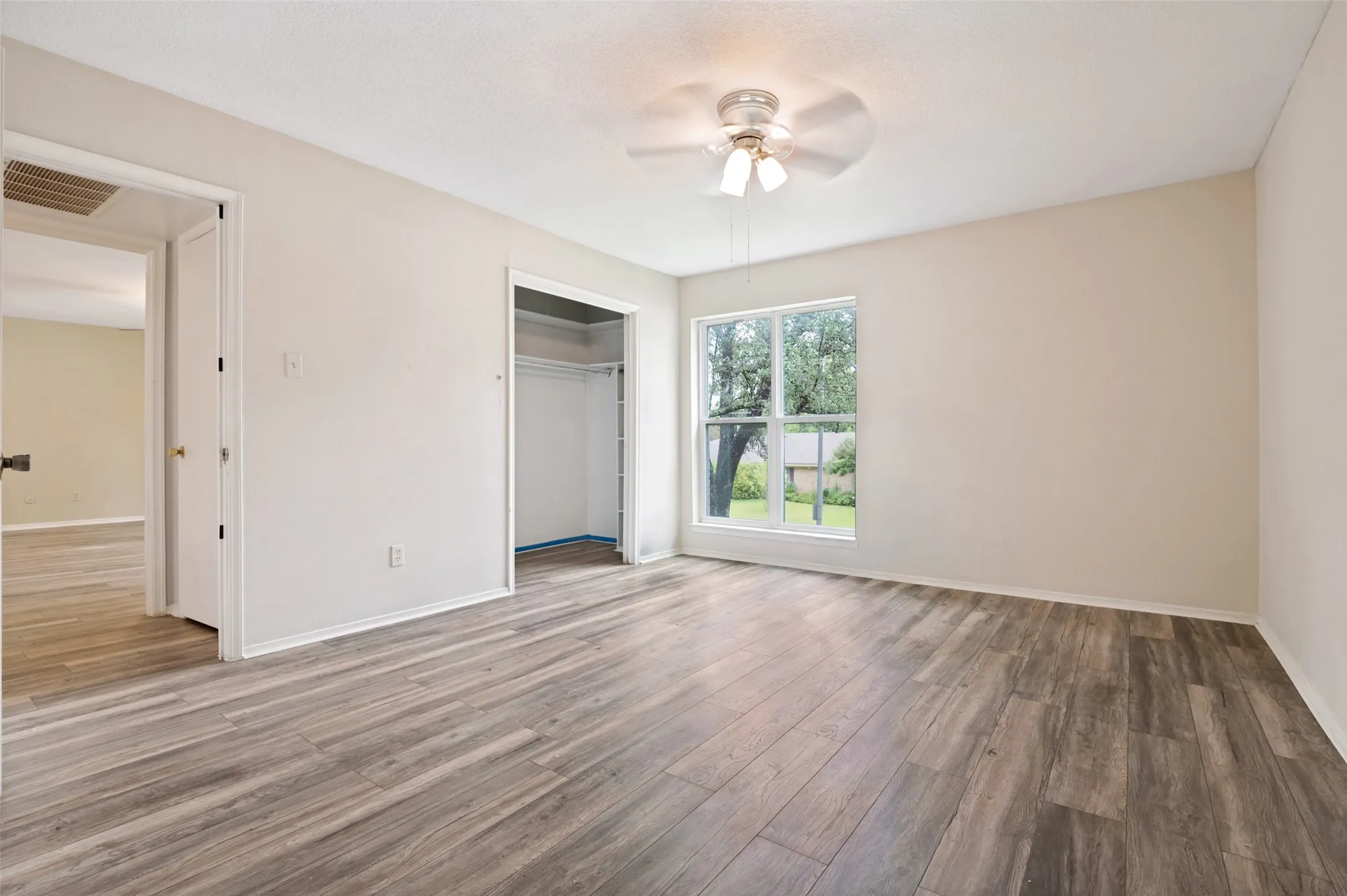 Spacious room featuring wood-finish flooring, a ceiling fan with integrated lighting, and a large window overlooking exterior greenery