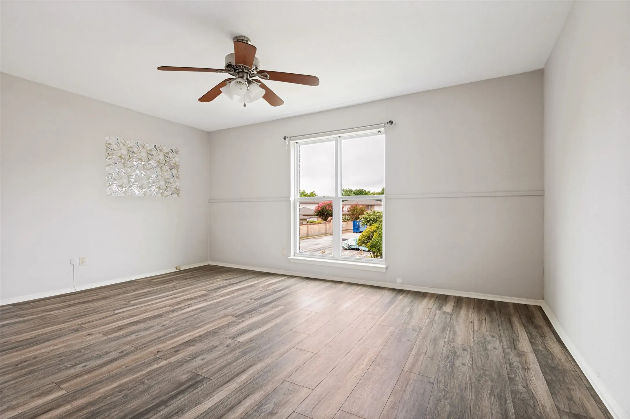Spacious room featuring wood-finish flooring, a large window, a ceiling fan with integrated lighting, and neutral wall tones