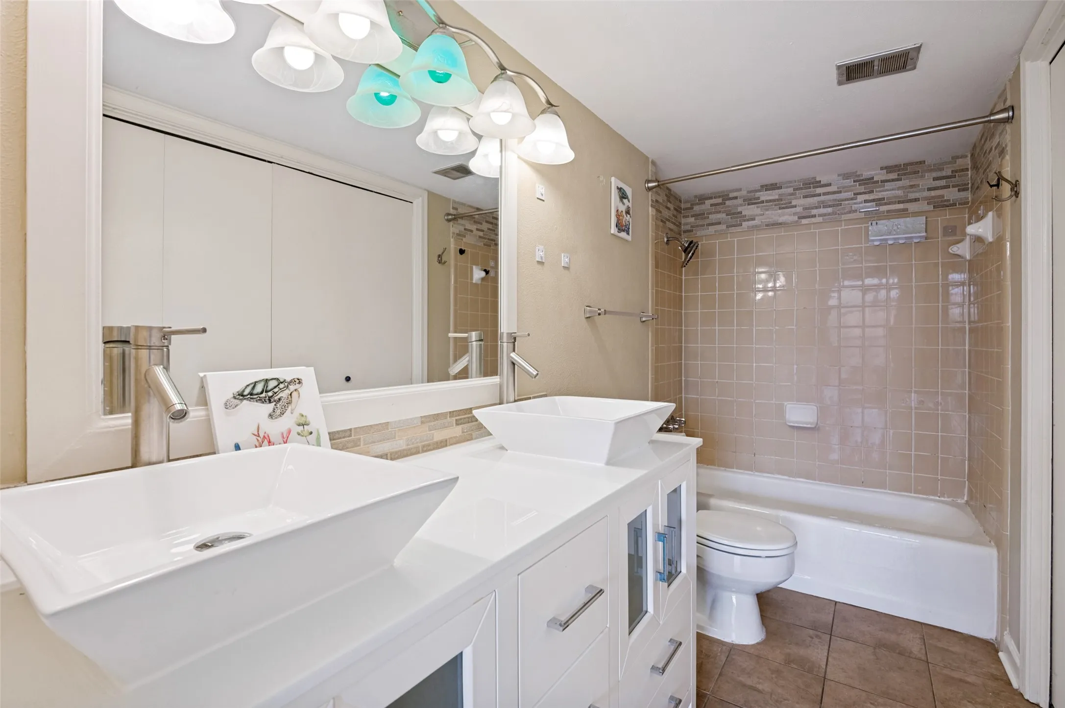 Dual vessel sinks with modern faucets, white vanity with cabinet storage, full vanity mirror, and a tiled shower/tub combination