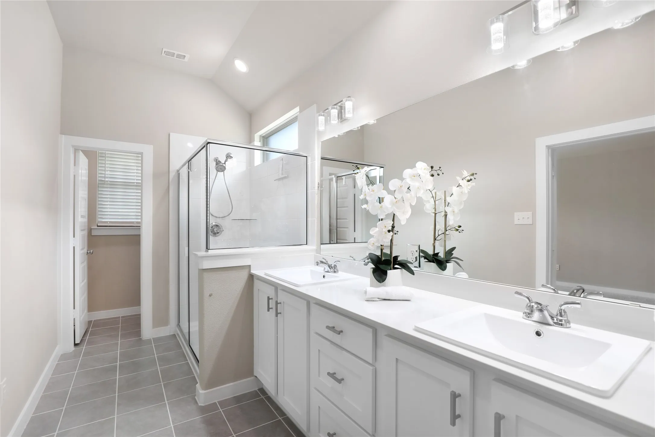 Spacious bathroom featuring a dual vanity with white cabinetry and integrated sinks, a walk-in glass-enclosed shower, tile flooring, and a vaulted ceiling