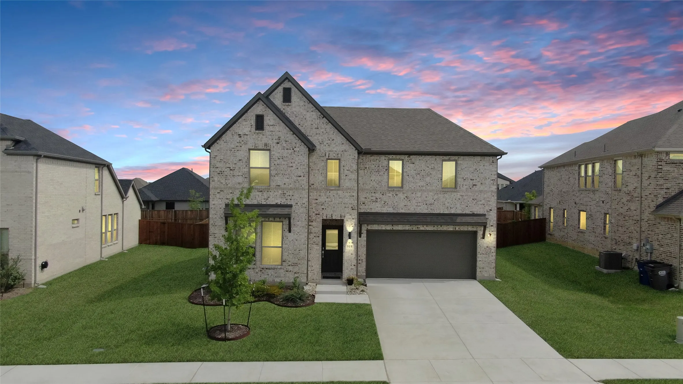 Two-story brick exterior featuring a dark gray roof, an attached garage, a concrete driveway, and a manicured lawn with a young tree