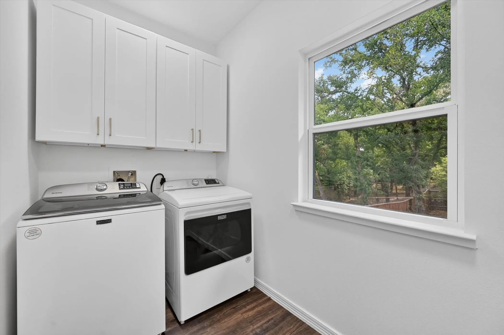 Laundry area featuring white shaker-style cabinetry with brushed nickel hardware, a large window, dark wood-finish flooring, and white walls