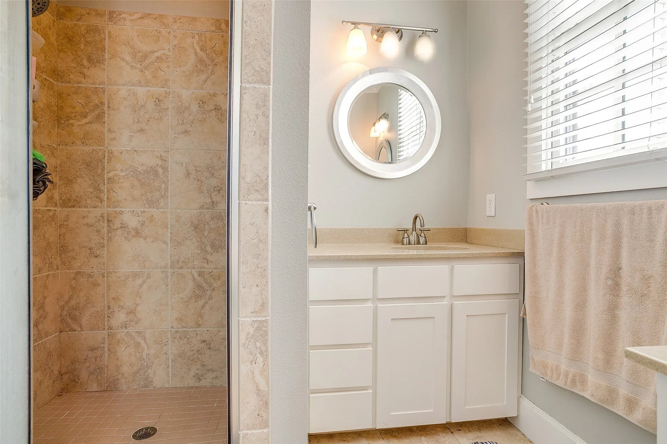 Bathroom featuring a tiled standing shower, white vanity with a light-toned countertop, round wall-mounted mirror, and a window with blinds