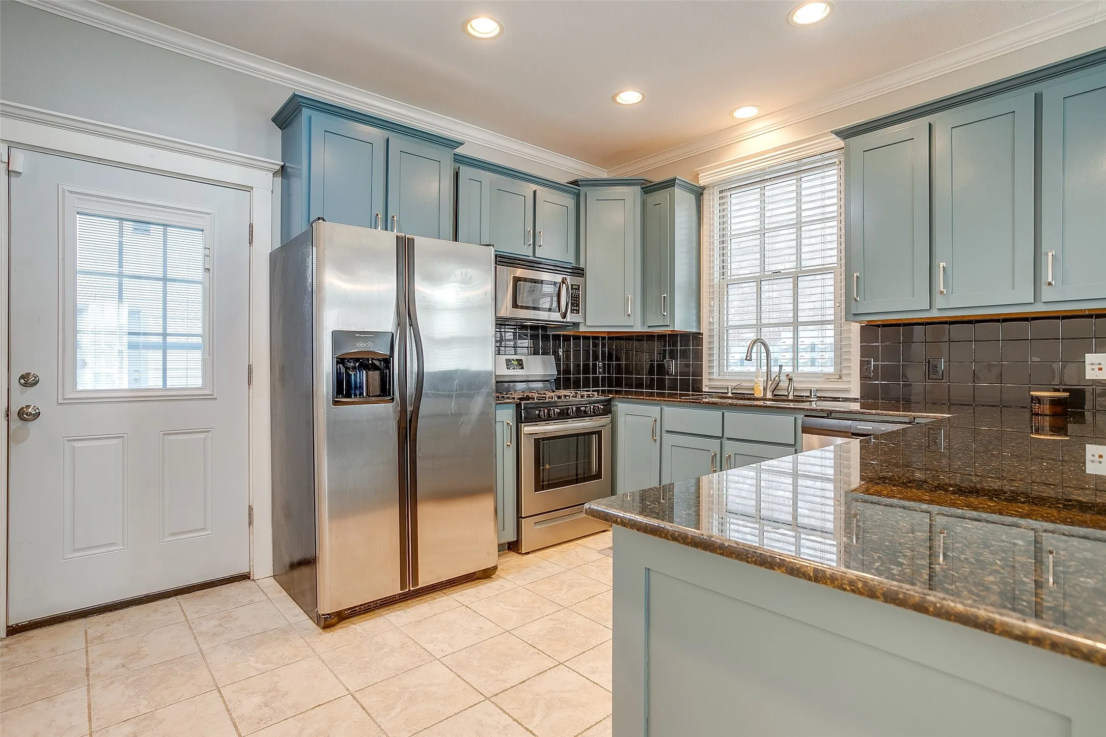 Kitchen featuring light blue cabinetry, dark granite countertops, stainless steel appliances, a dark tiled backsplash, and recessed lighting